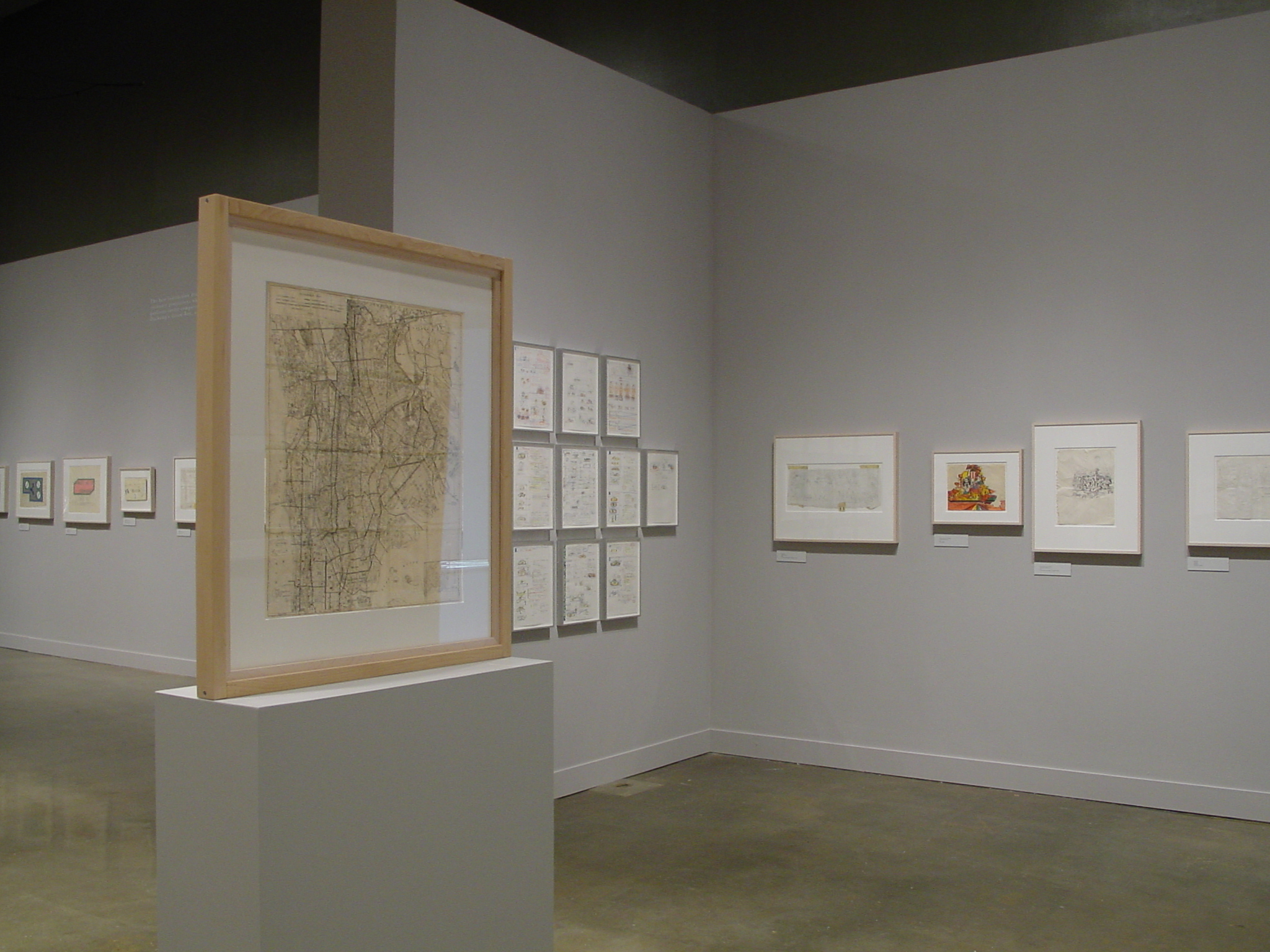 On a pedestal, a wooden frame houses a drawing on yellow paper. Gallery walls are filled with additional framed drawings