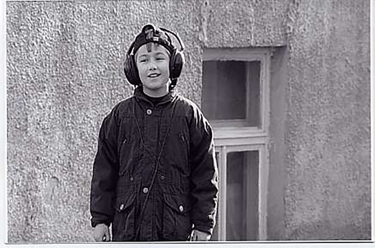 Black and white video still of a young boy wearing headphones over his ears, in front of a concrete building