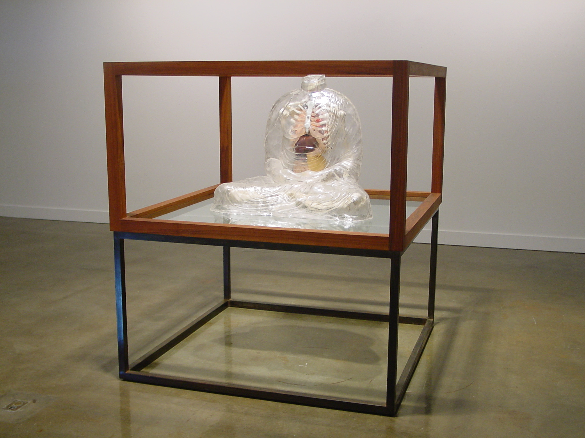 A three dimensional work resembling a human form sits cross legged in an elevated framed stand