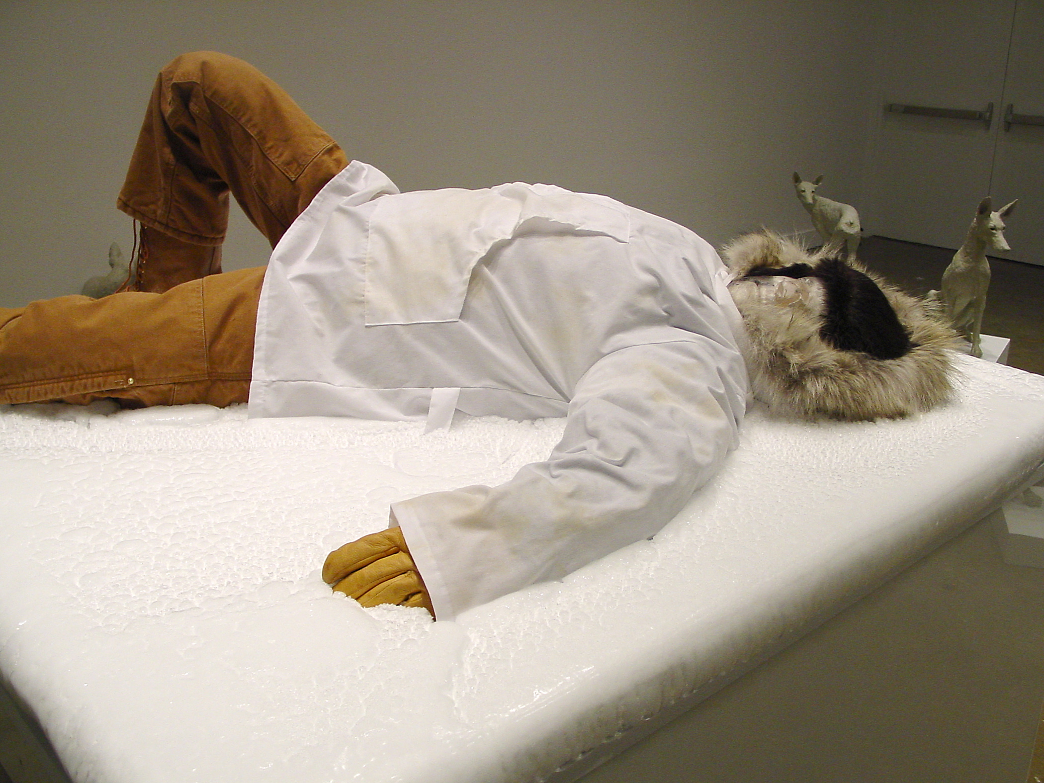 Close up view human-like artwork, lying on a tall white pedestal, clothed in winter attire with a fur hood and boots