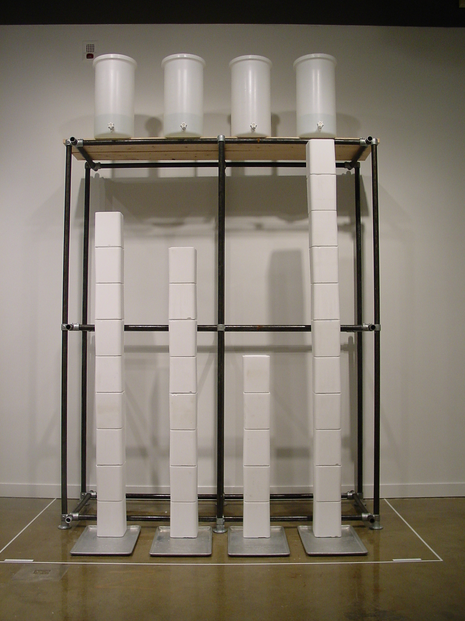 White compressed salt blocks stack top 4 aluminum trays. Behind, a black framed structure is against the wall