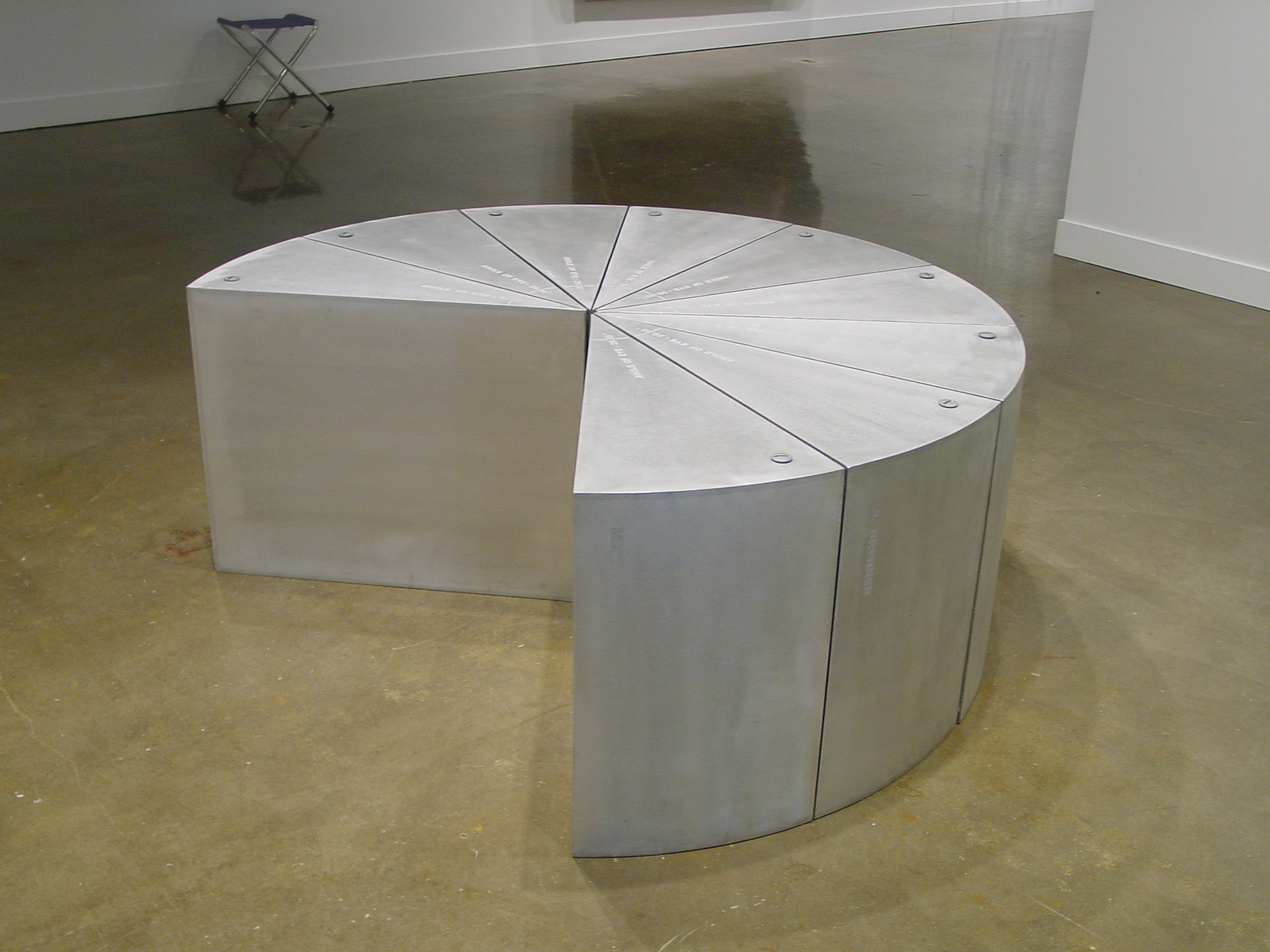 An aluminum sculpture on gallery floor similar to a 3 dimensional pie chart, with missing pieces