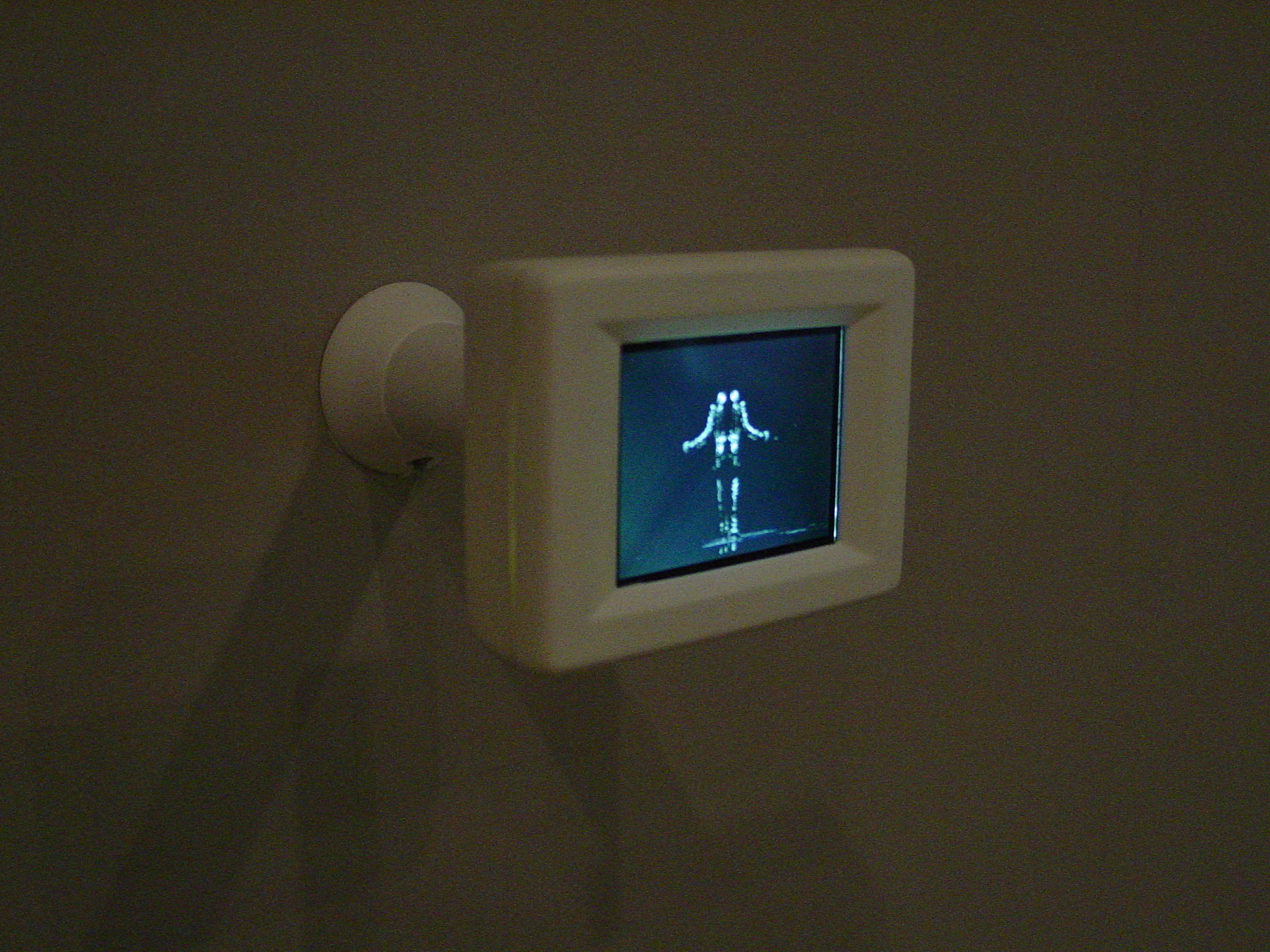 Small, white framed digital screen is mounted to the wall, displaying a white human-like form on a black background