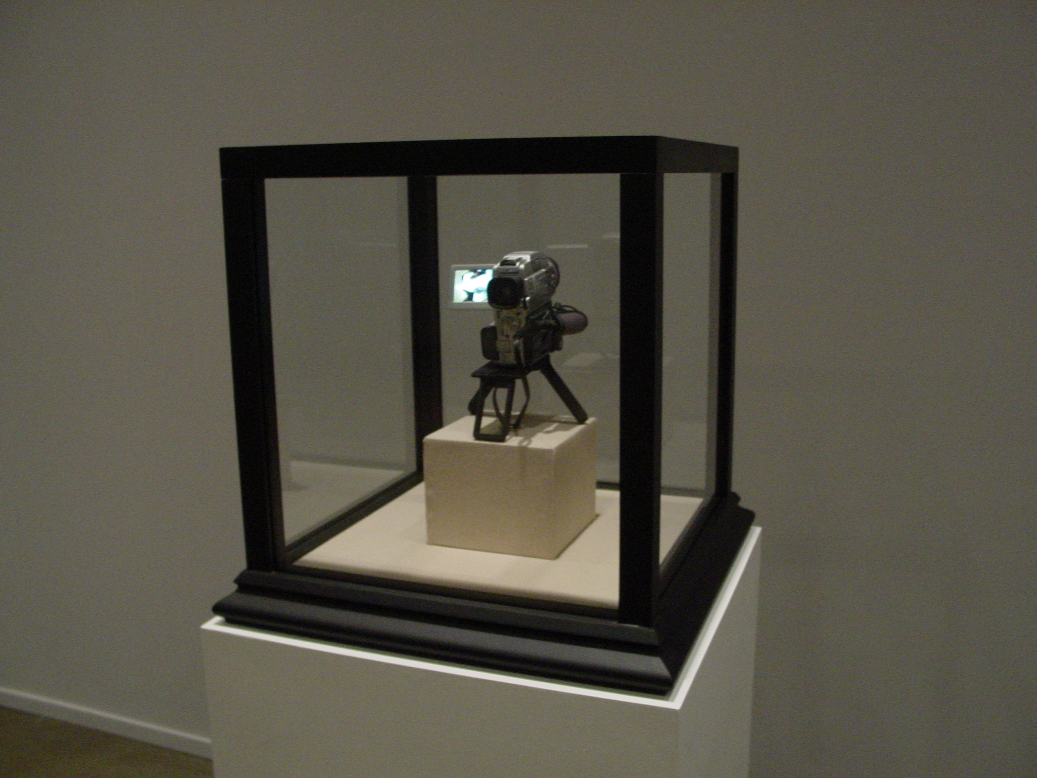 A digital camcorder sits on a pedestal within a vitrine, framed with black edges.
