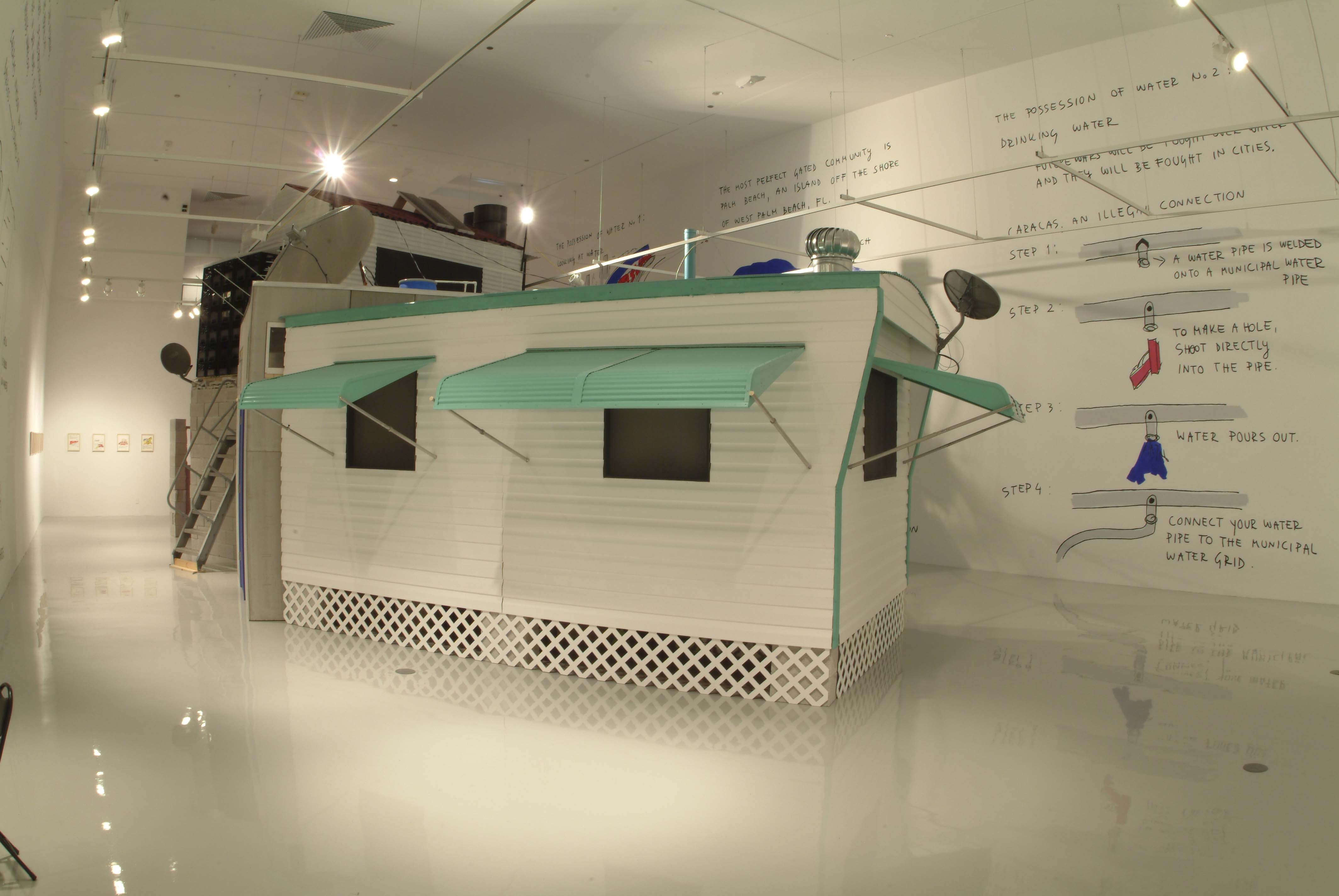 A small white house with turquoise awnings sits in gallery space with writings on the wall behind