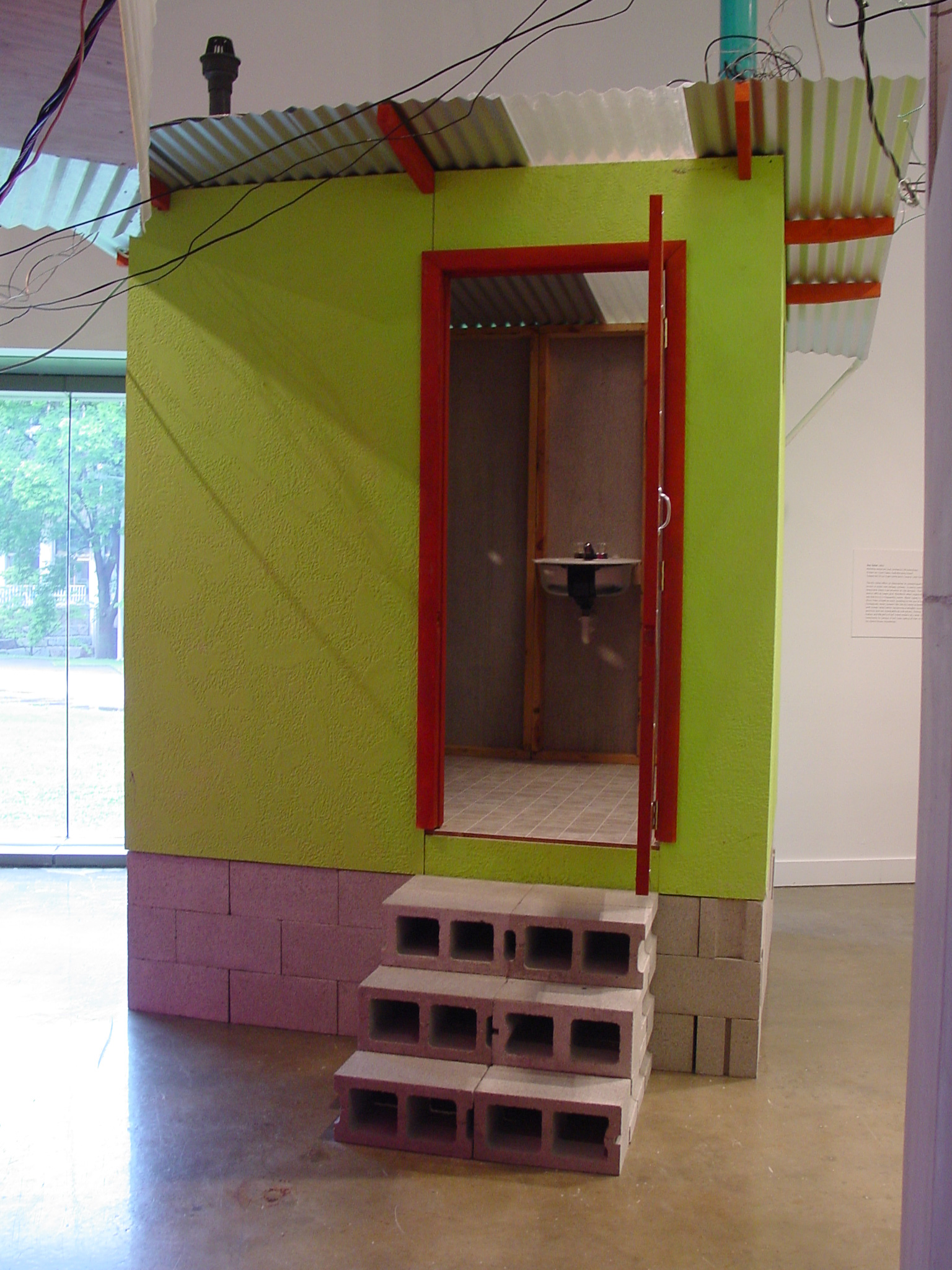A lime green out house with a metal roof and an open red doorway is raised on cinder blocks