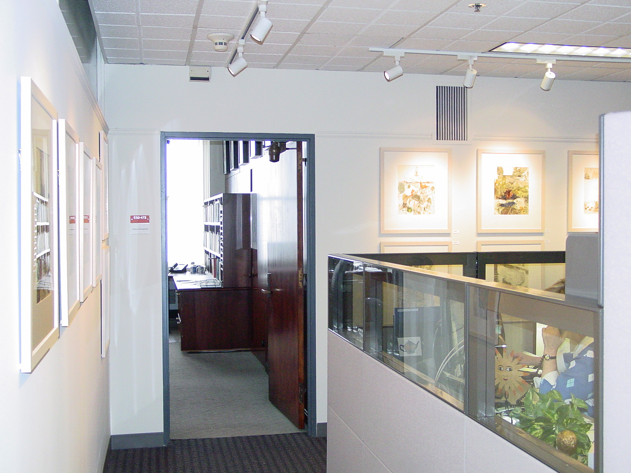 A view into an office from a hallway where many brightly lit framed abstract artworks hang in grids across from cubicles.