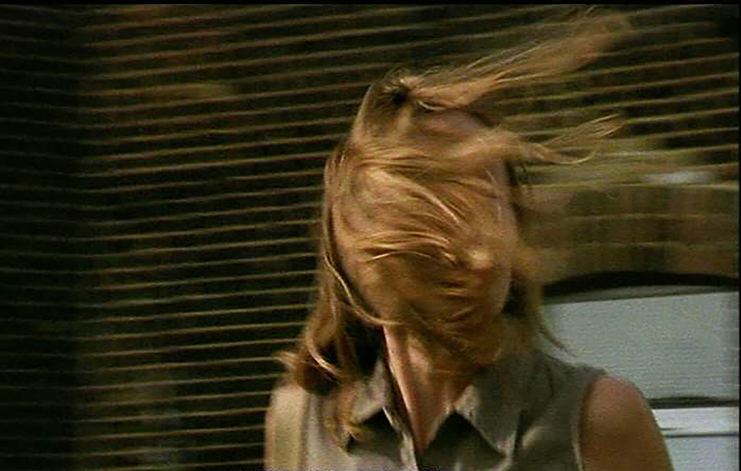Video still freezes on a woman, in front of a brick wall, with wind blown straight blonde hair covering her face