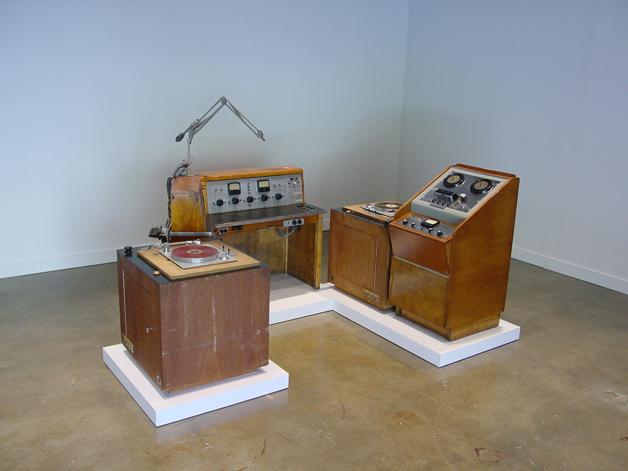 Vintage wood paneled radio broadcast equipment installed on low white pedestals on the floor in the corner of the gallery.