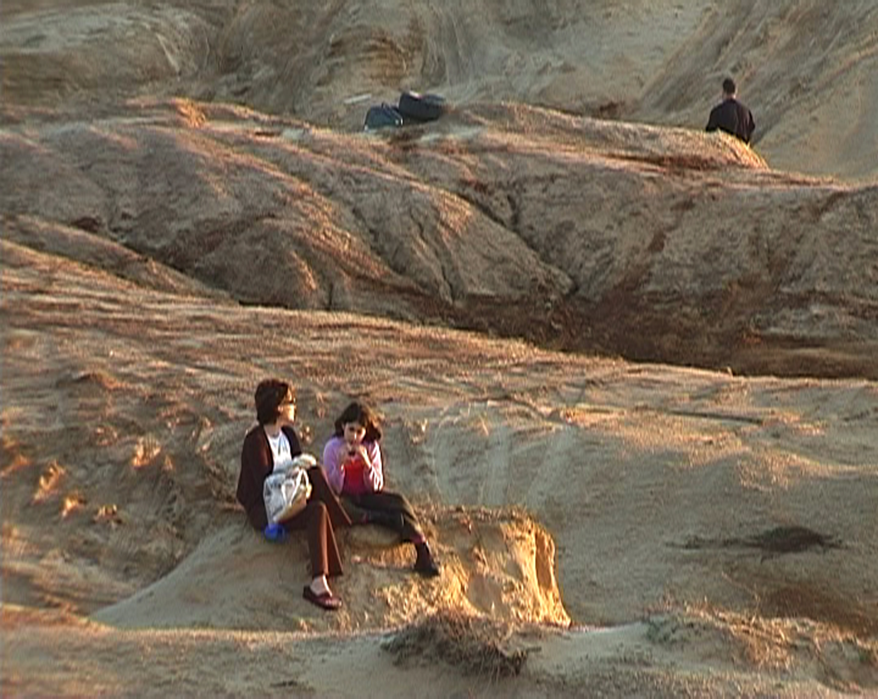 A woman and child sit on a sandy coastal hill outside of Tel Aviv, near an area where men drive trucks off road.