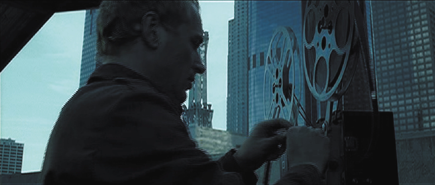 Wide panoramic film still showing a man with his hands on a reel to reel film projector with a cityscape in background.