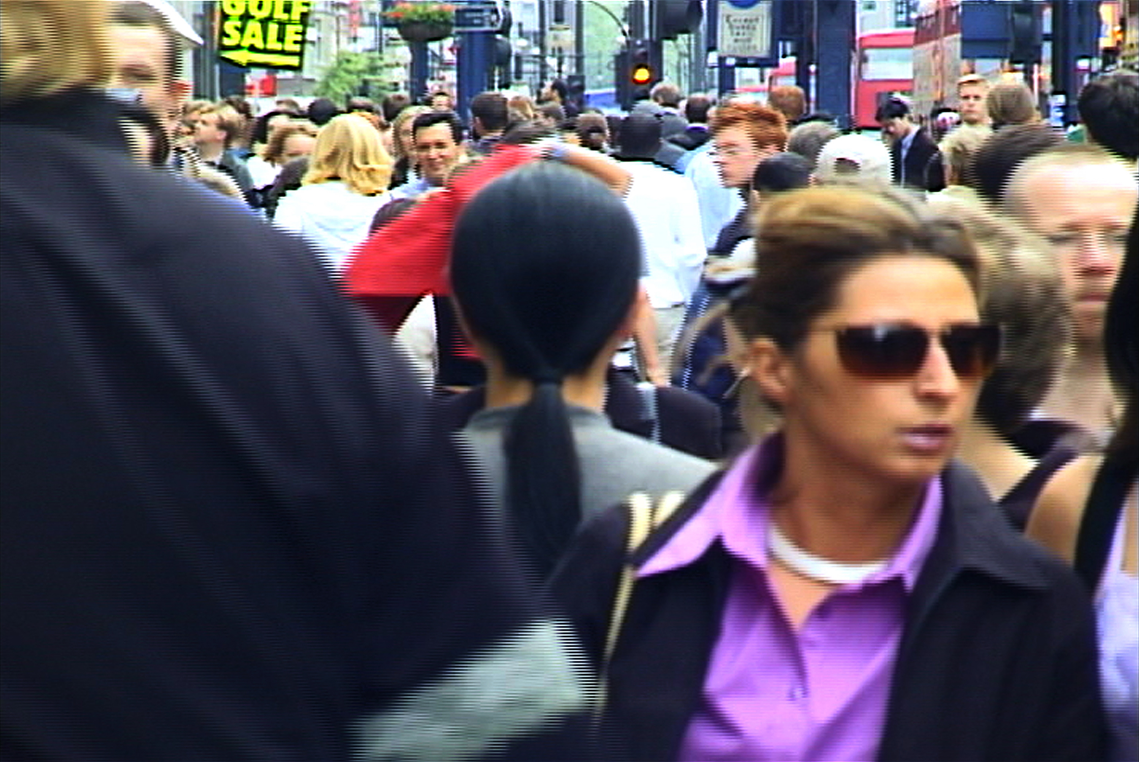 A woman with long black hair dressed in gray clothing stands with her back to the camera on a crowded street in London. 