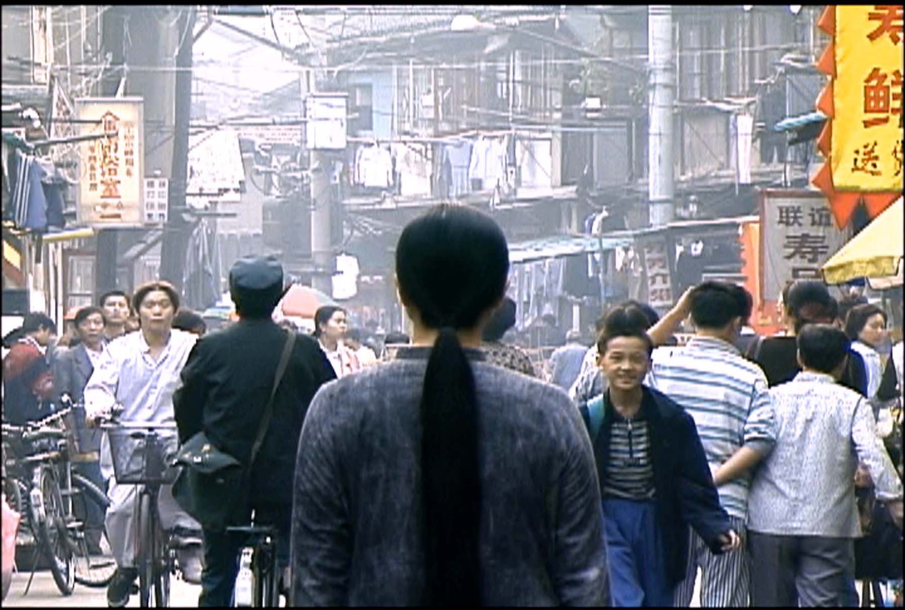 A woman with long black hair dressed in gray clothing stands with her back to the camera on a crowded street in Shanghai.