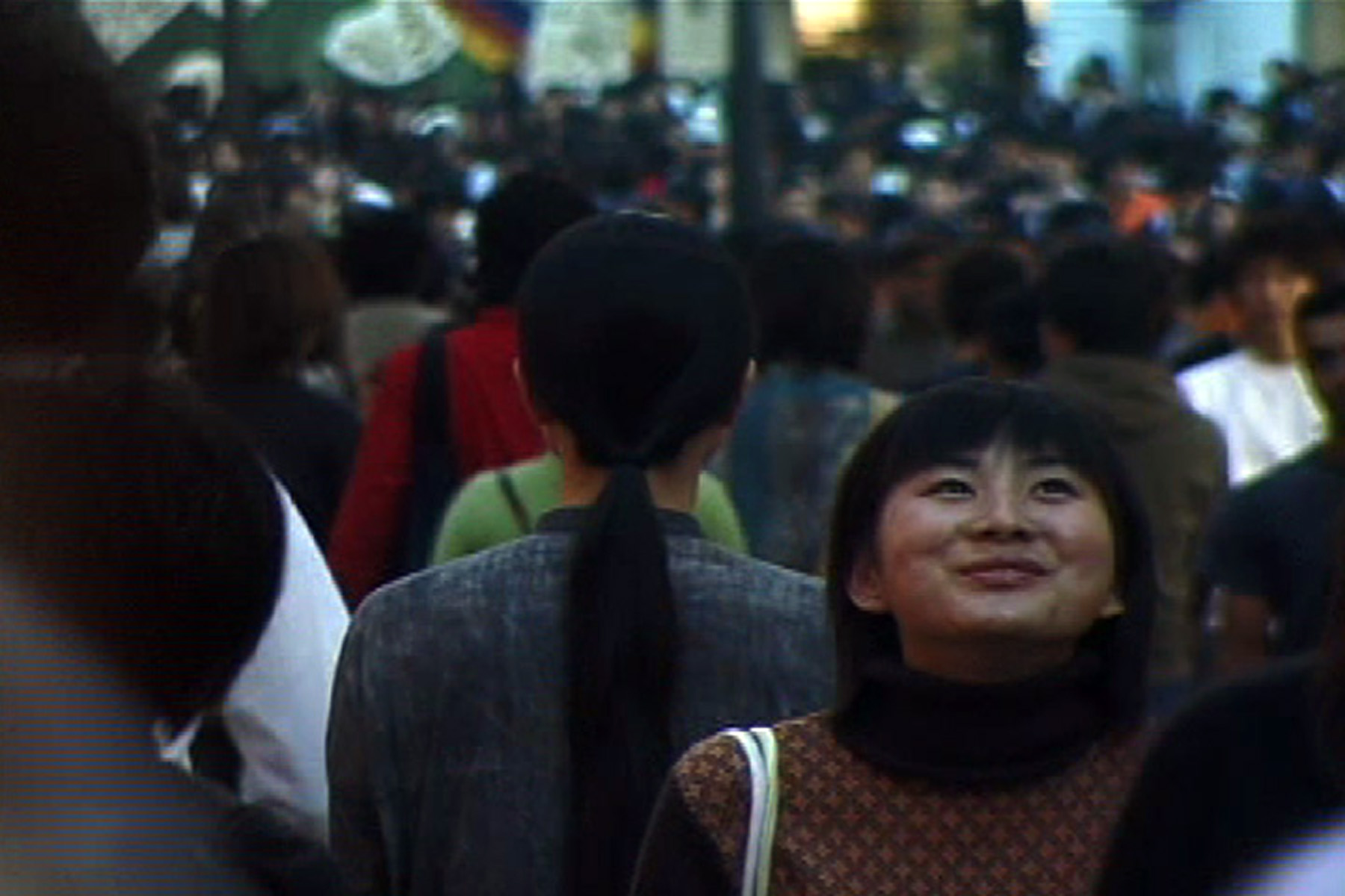 A woman with long black hair dressed in gray clothing stands with her back to the camera on a crowded street in Tokyo.