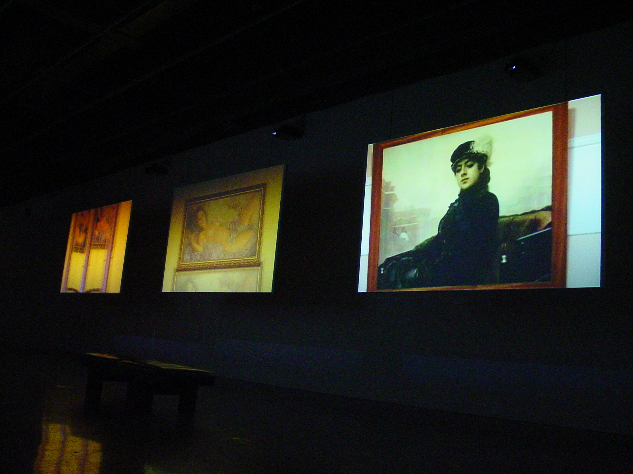 A darkened room with a bench in front of three large video projection screens showing color images, one is a woman’s portrait.