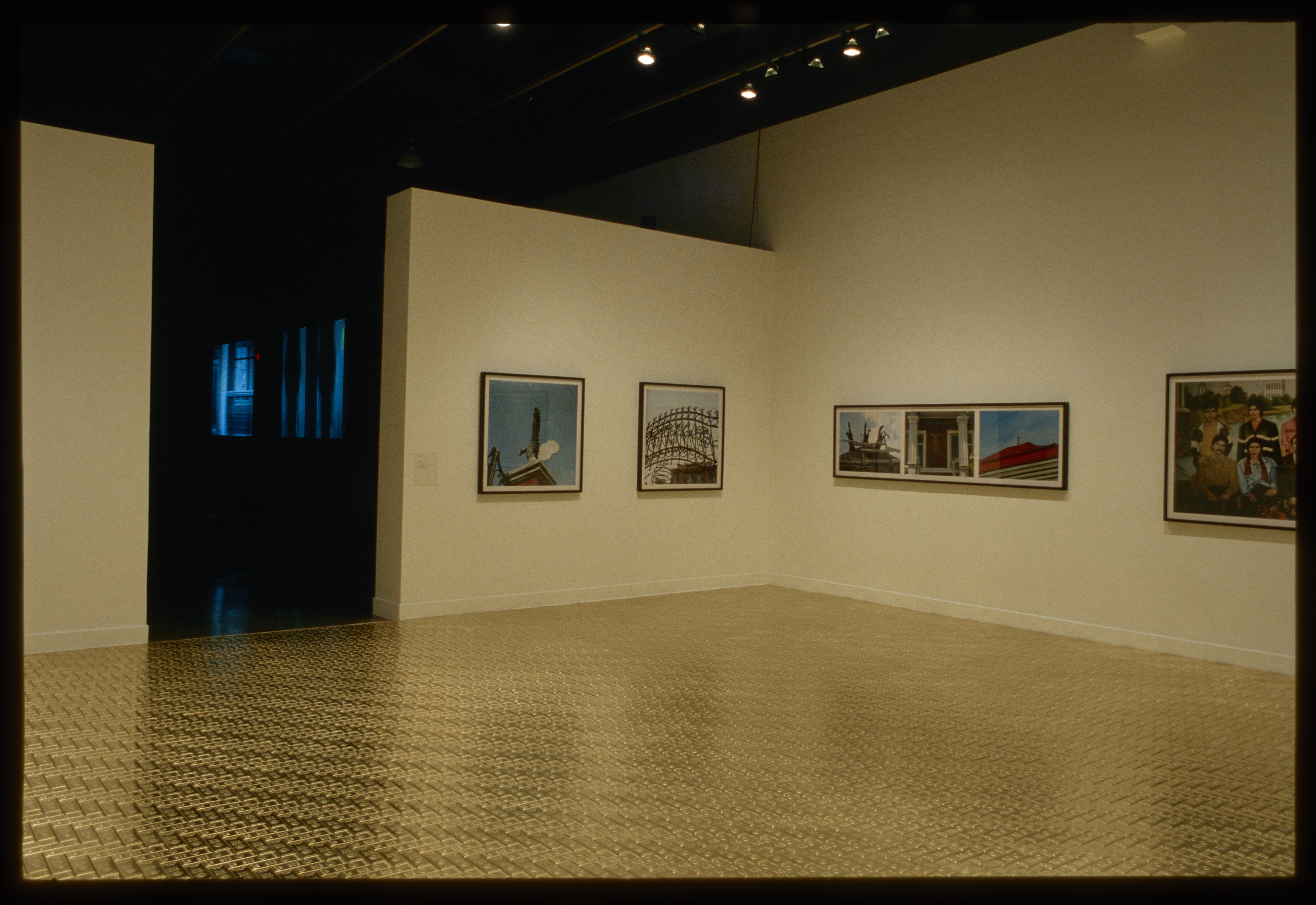 A room with a shiny golden floor and large colorful photos hung on two walls leads to a darkened room with projected videos.