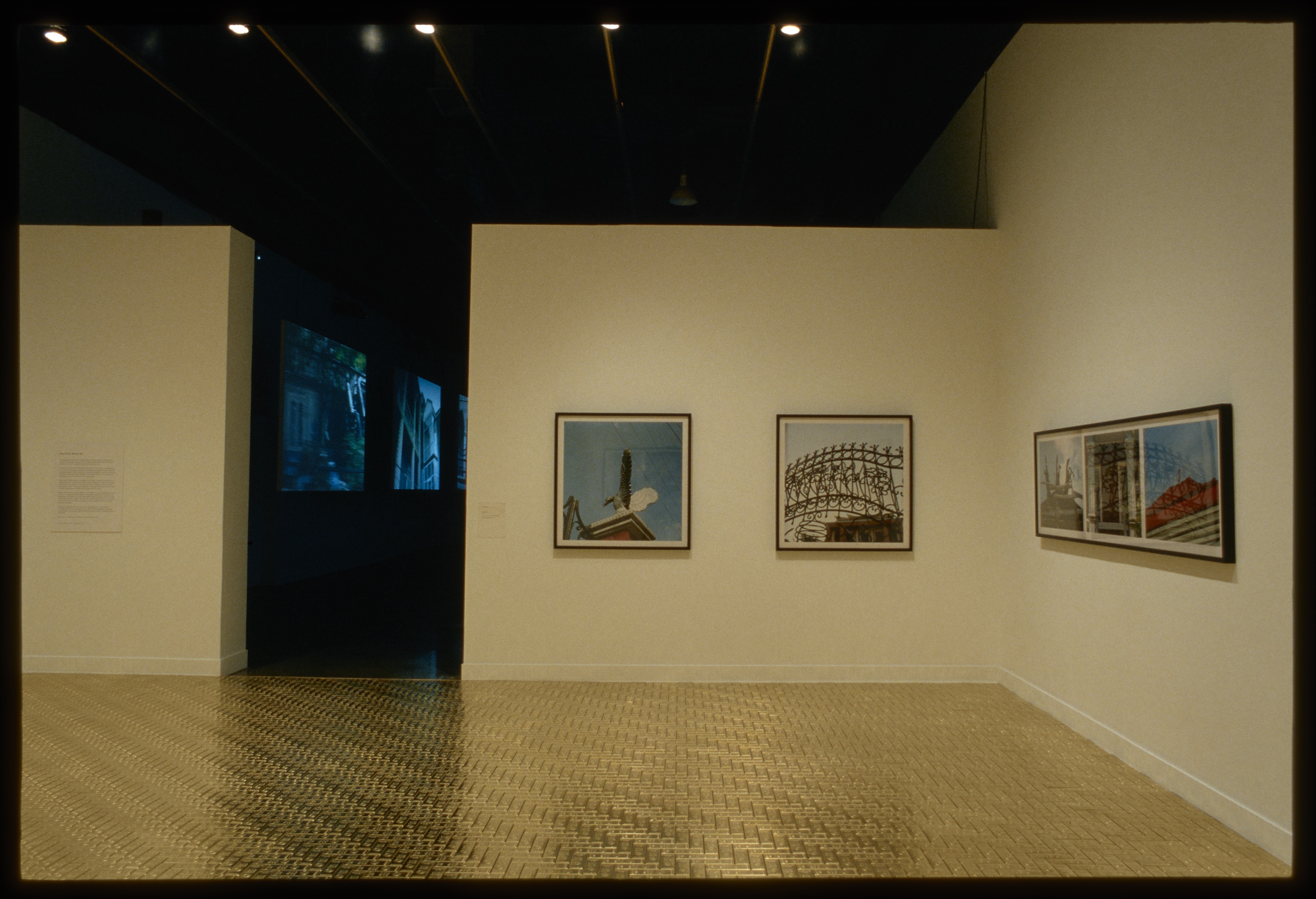 A room with a shiny golden floor and large colorful photos hung on walls leads to a darkened room with projected videos.