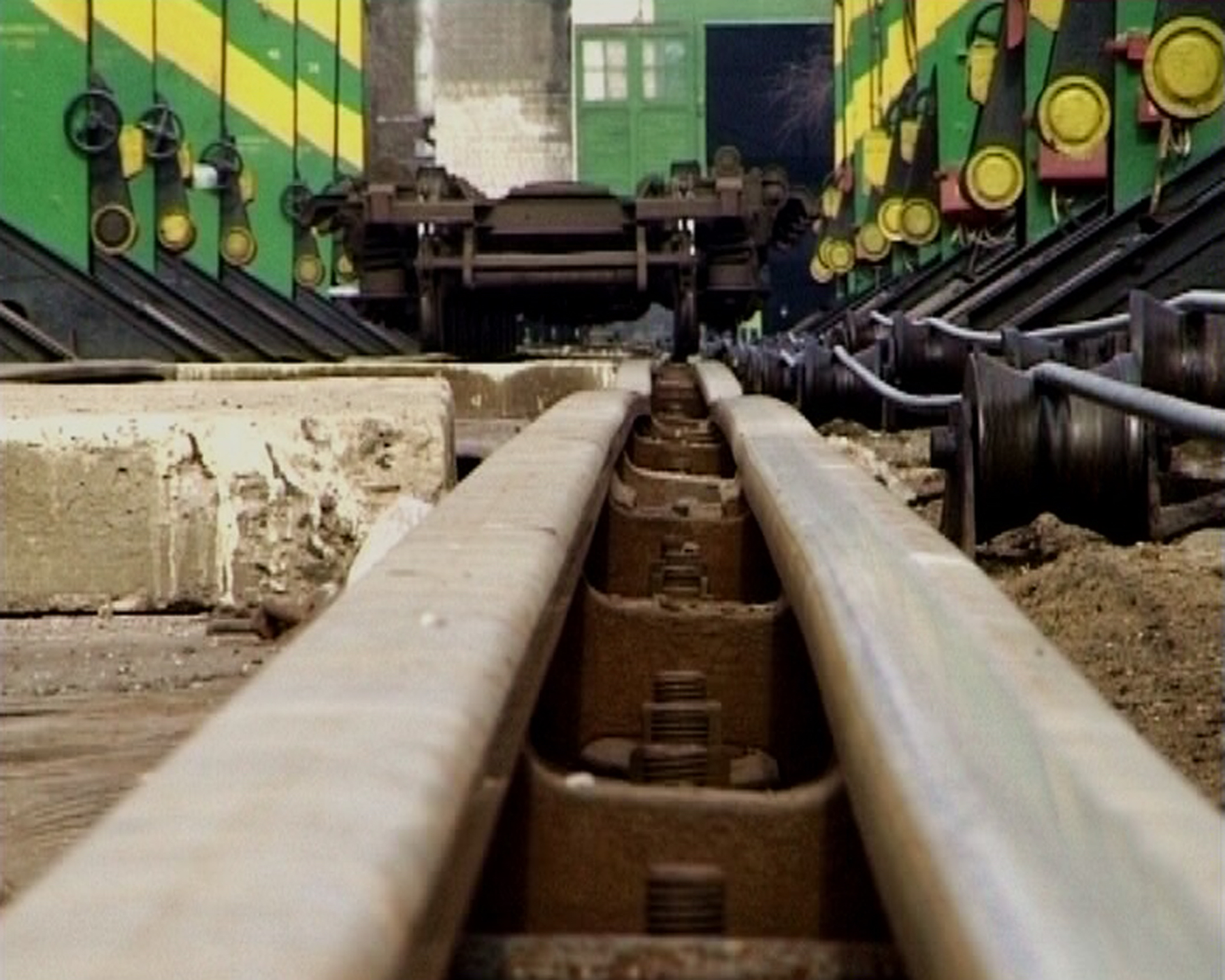 Video still showing a close view of a track for a train car in the foreground, and a train car chassis in the background.
