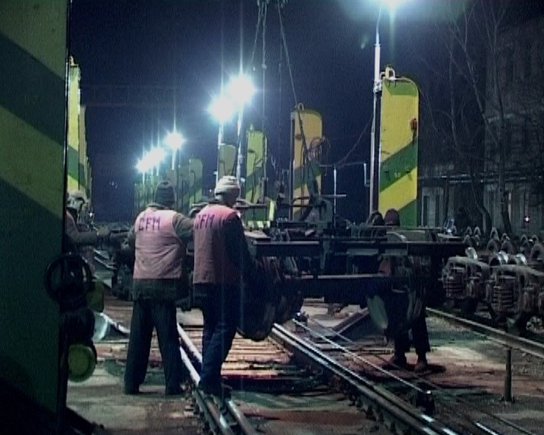 Video still showing workers in an industrial station with bright painted pillars lifting a train chassis above train tracks. 