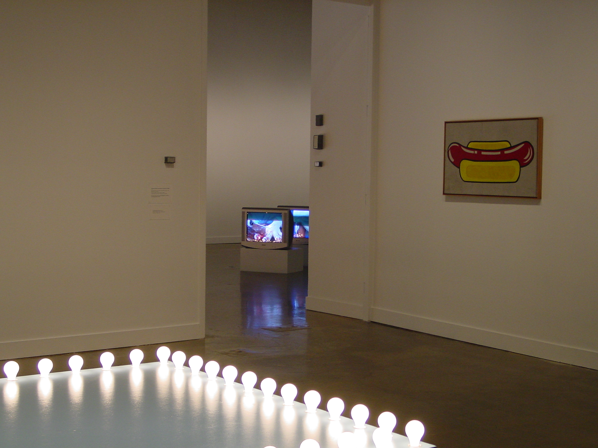 Platform stage adorned with ball lights, a painting of a hotdog on wall, and videos playing on tv monitors in the background.