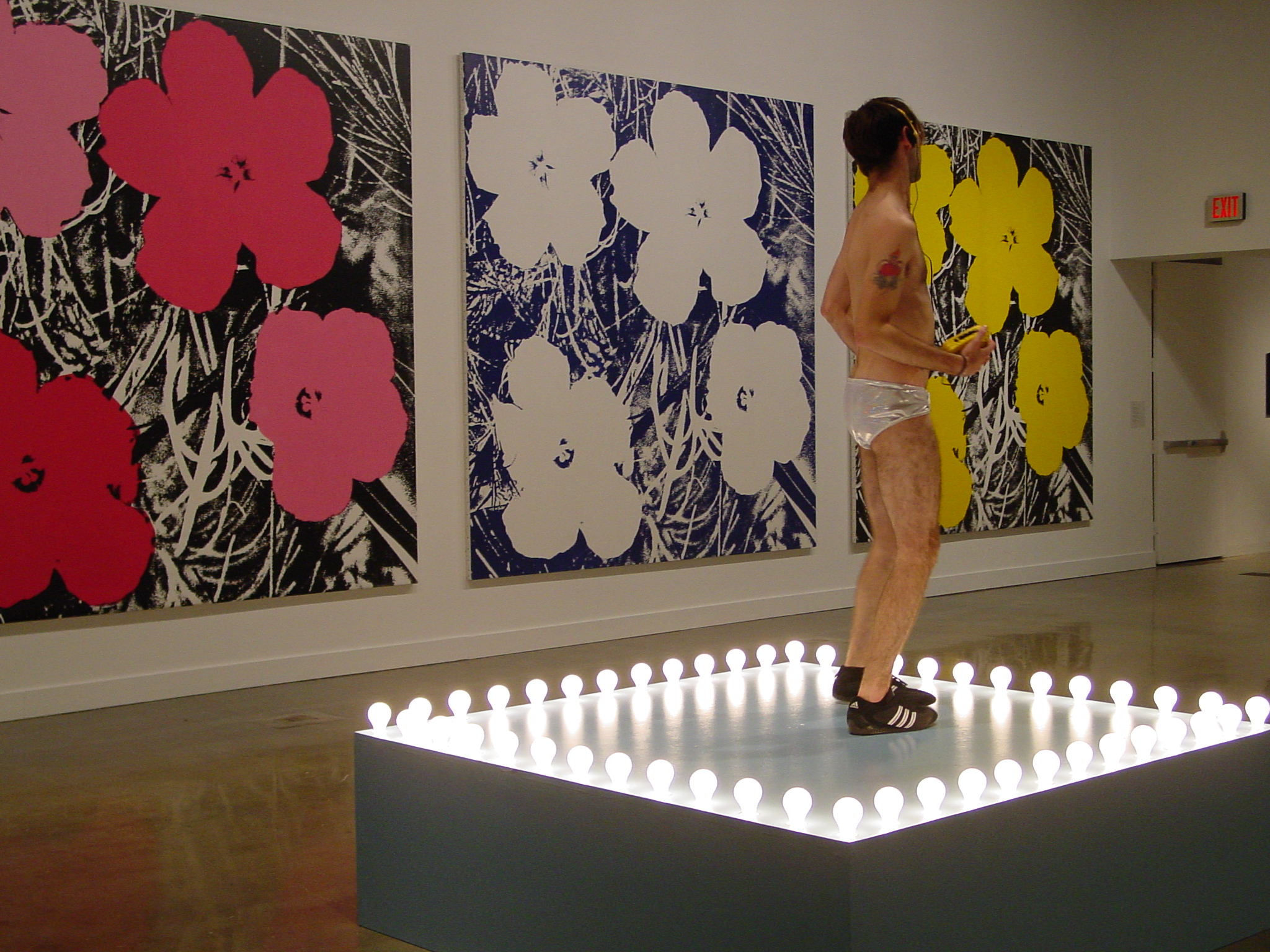 Male dancer on lit stage in a speedo, shoes, listening to a yellow walkman, with hot dog painting and video monitors behind.