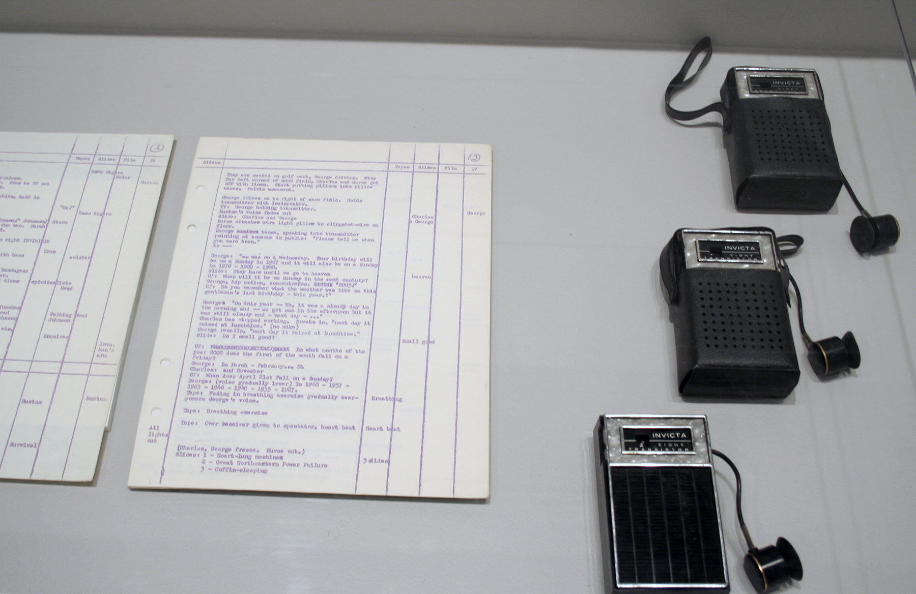 Close view of two handwritten documents and three small vintage hand crank radios arranged under a clear case on a pedestal.