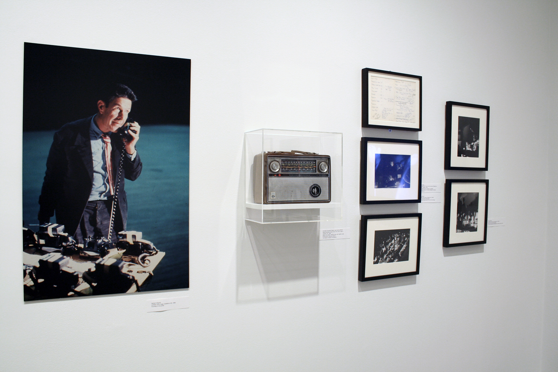 Large color photo of man in suit holding telephone receiver, a radio on a shelf in a case, small framed pieces hung on the wall.