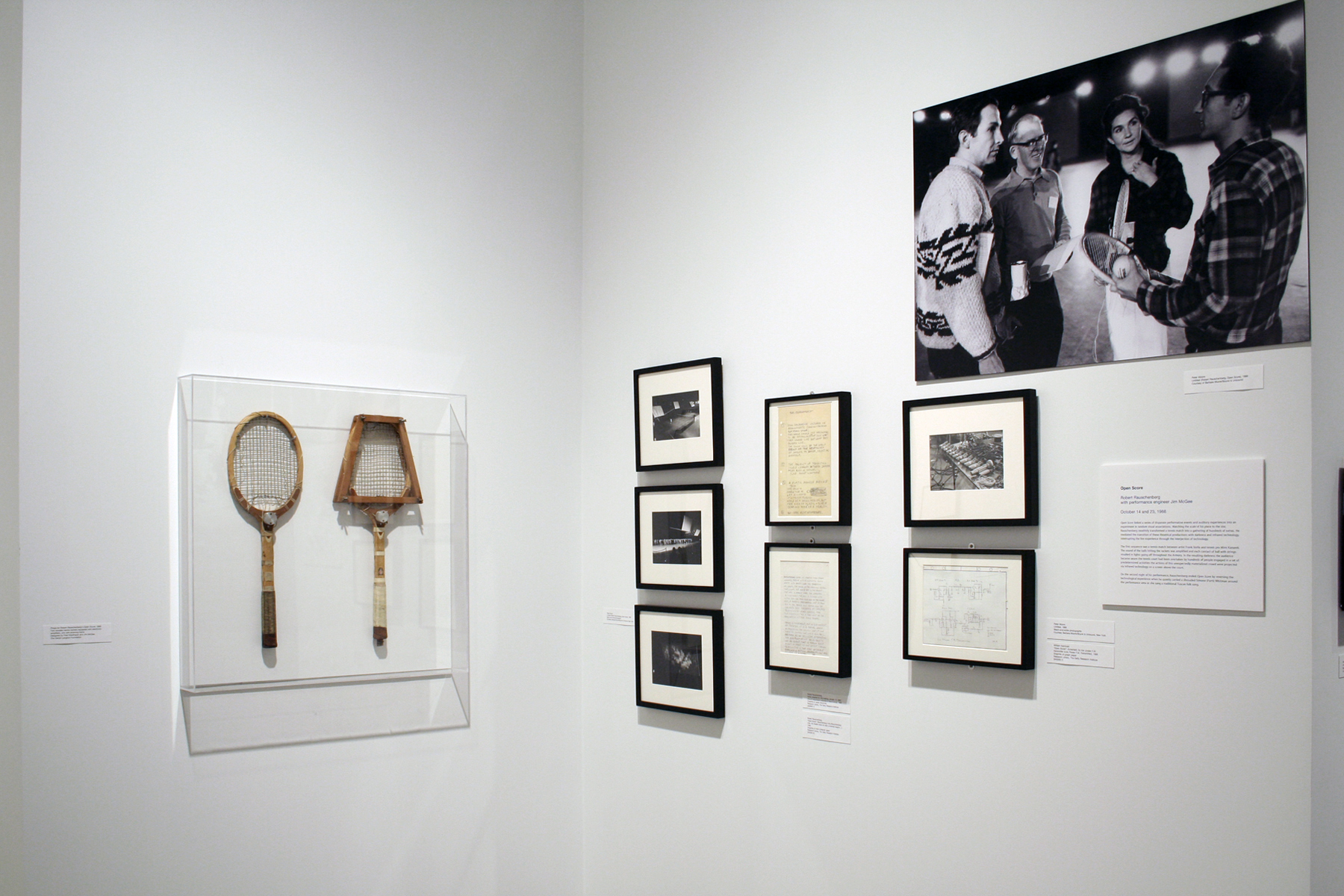 Two tennis rackets in a clear case, a group of small photos, documents, and a large black and white photo hang on two walls.
