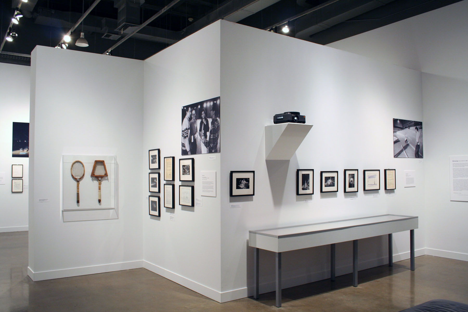 View of gallery shows 2 tennis rackets in a case, framed works and large photos mounted on walls and a glass top display case.