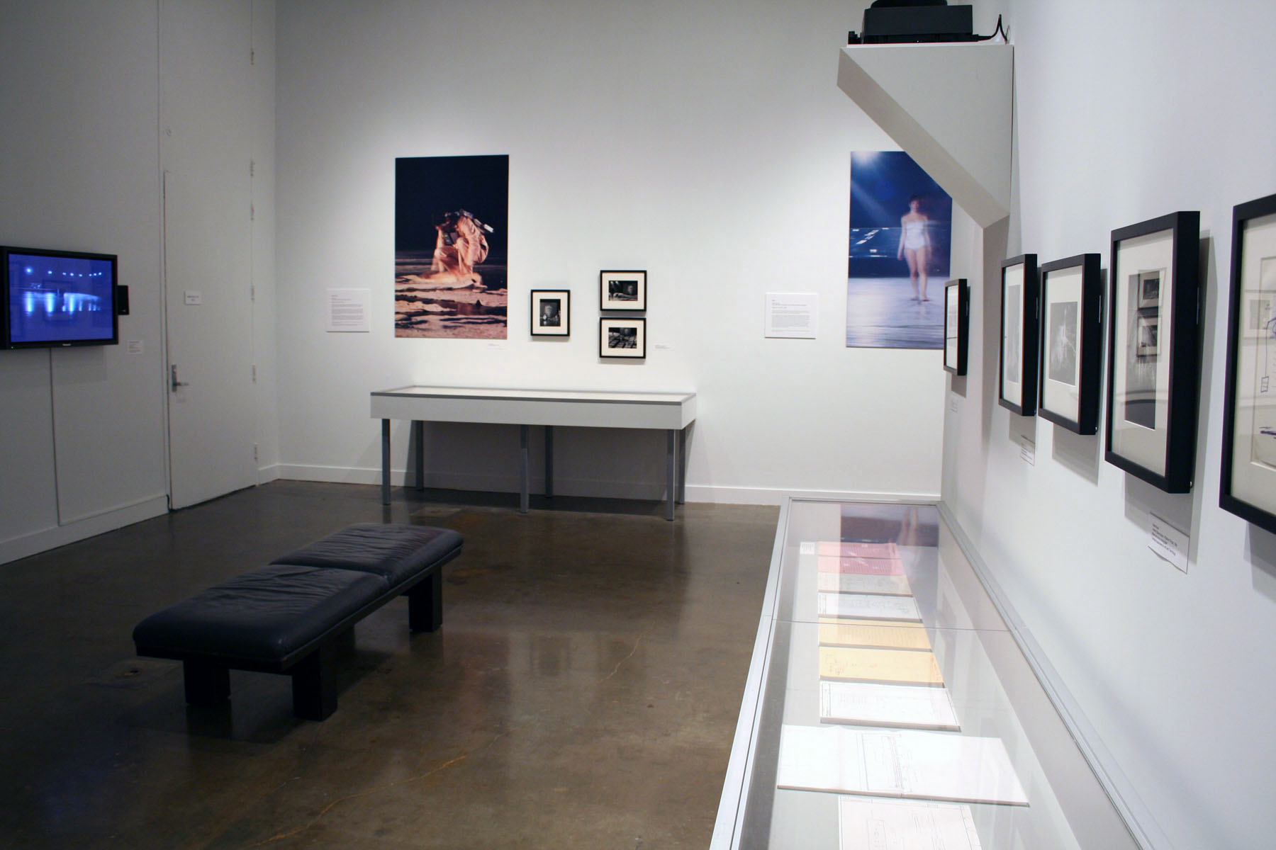 Miscellaneous framed photographs hang against a white gallery wall while paper artifacts sit in a glass vitrine.