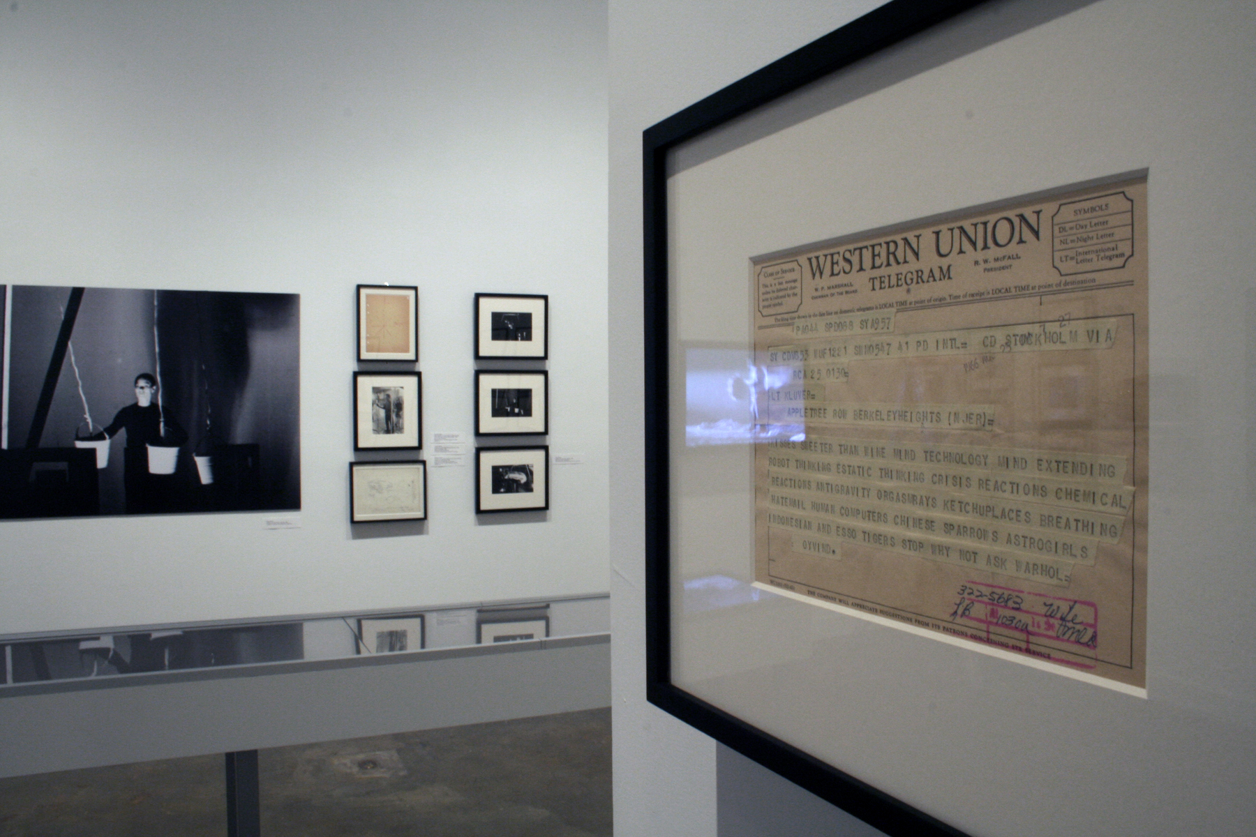 View of gallery with framed artworks and photos hung on walls, a long display case, and a video monitor with a bench in front.