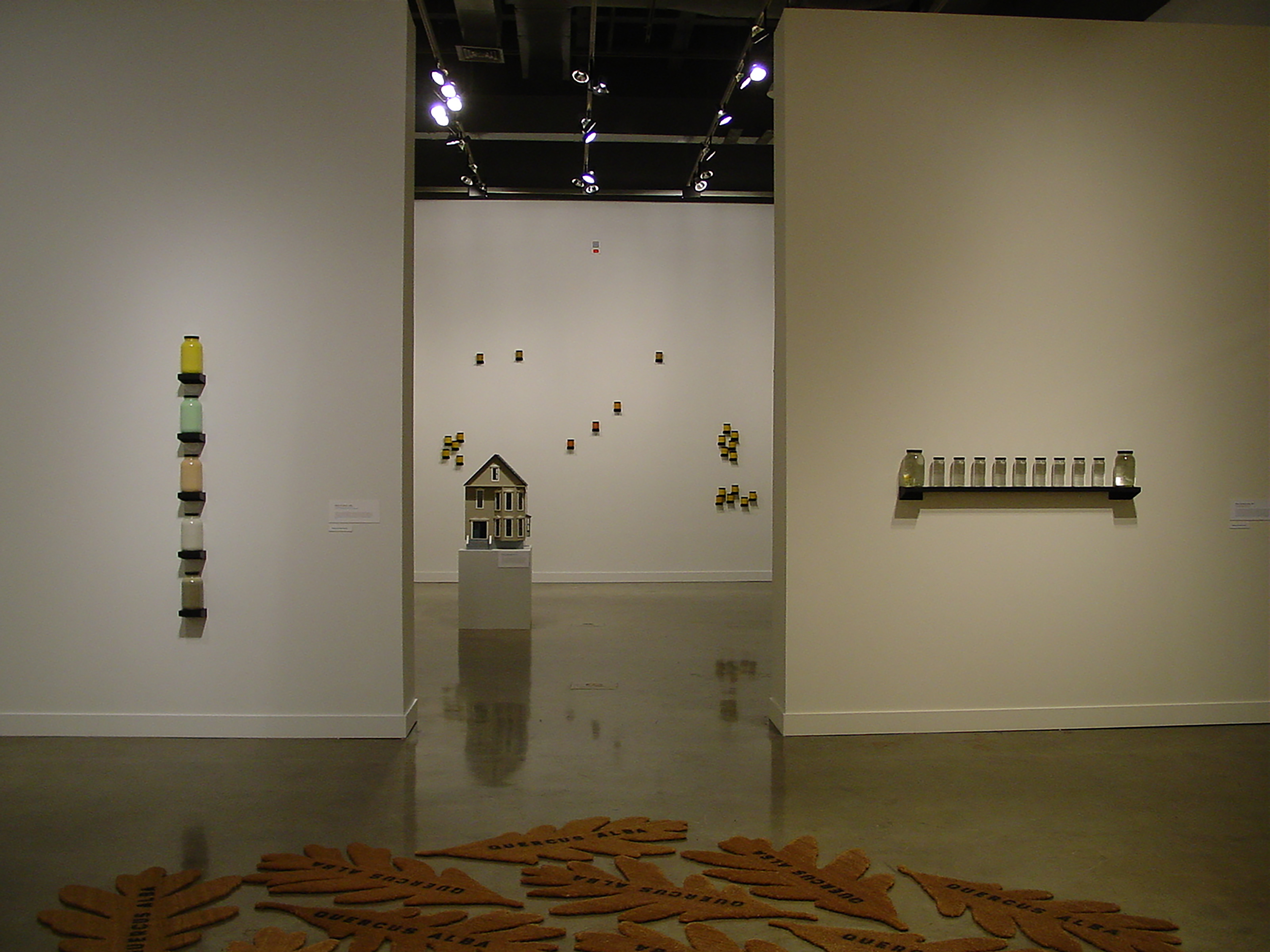 Oak leaf shaped mats lead to glass jar artworks on walls, a drab model house on pedestal and artworks hung in the back room.