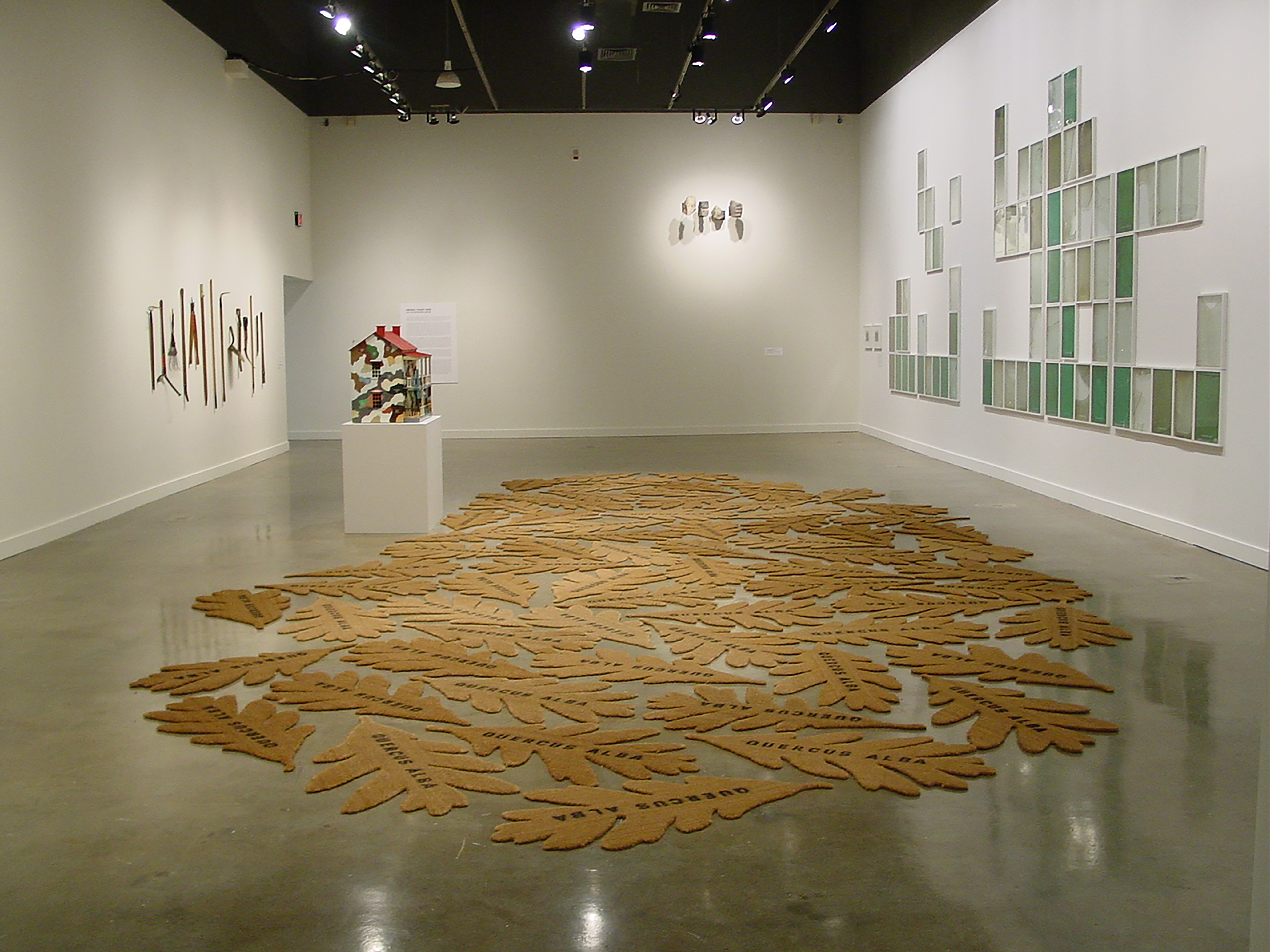 Multitude of oak leaf shaped mats beside a colorful model house sculpture on pedestal and 3 artworks on surrounding walls.