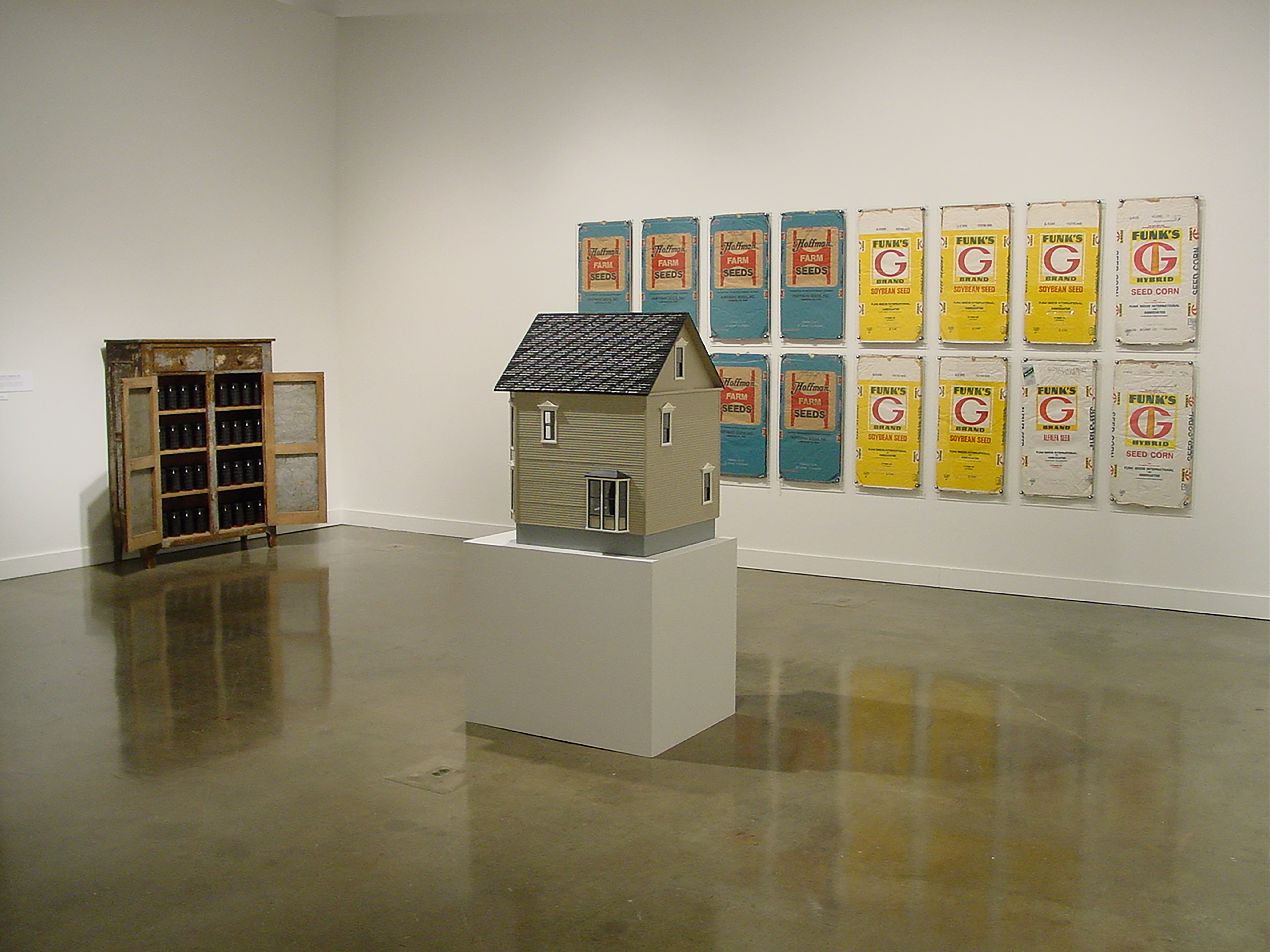 A drab colored model house on a pedestal, a cabinet with open doors to the left, and a group of bright artworks hang behind.