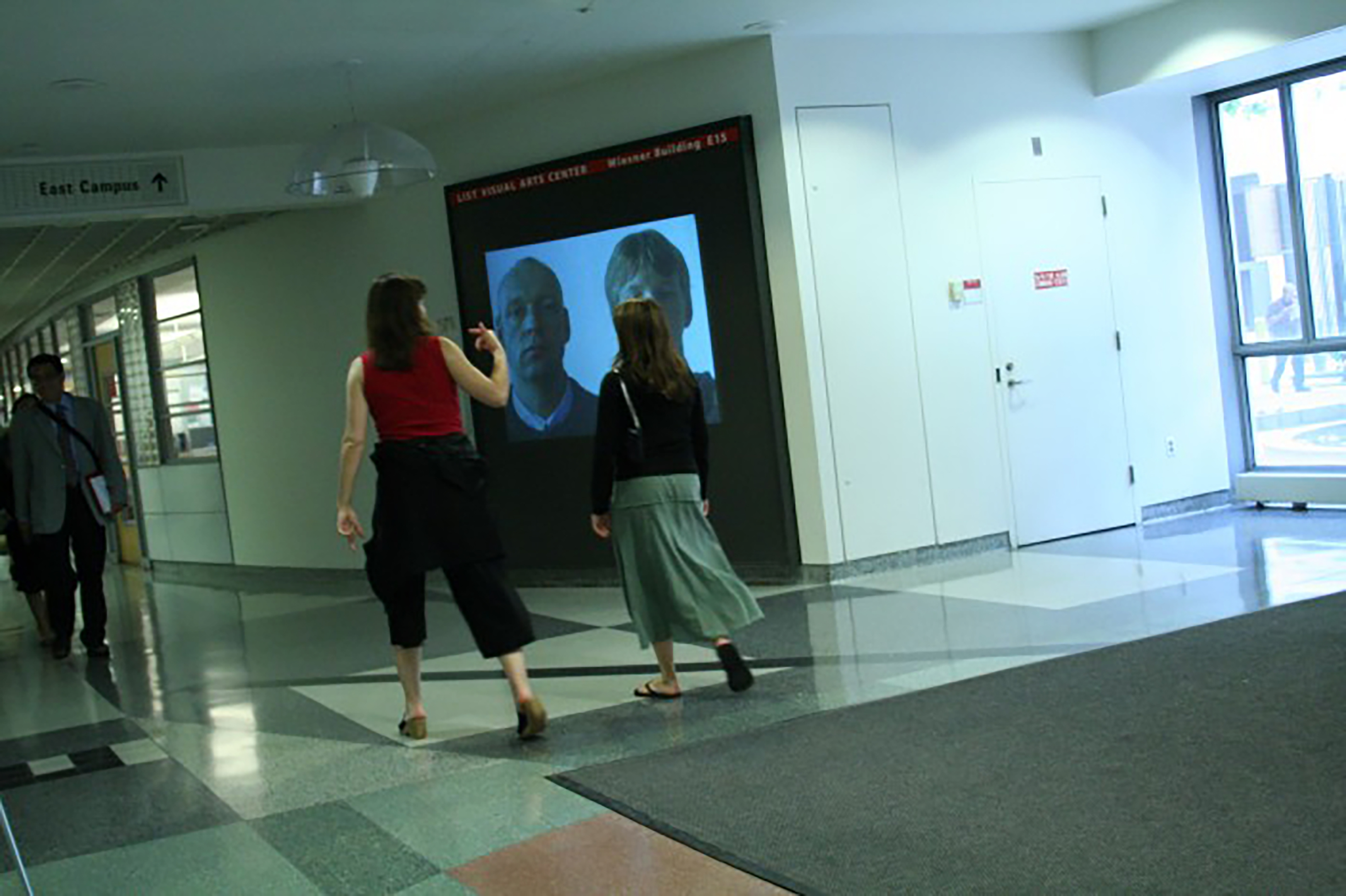 A view of a projection space in a hallway at MIT showing a portrait of the artist and man side by side.