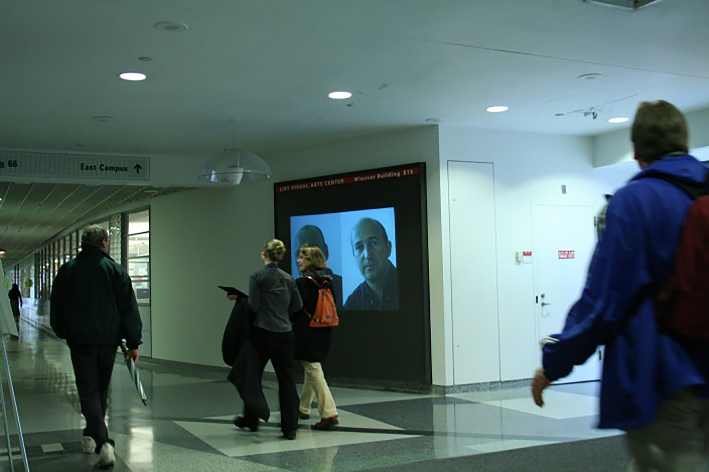 A view of a projection space in a hallway at MIT showing a portrait of the artist and man side by side.