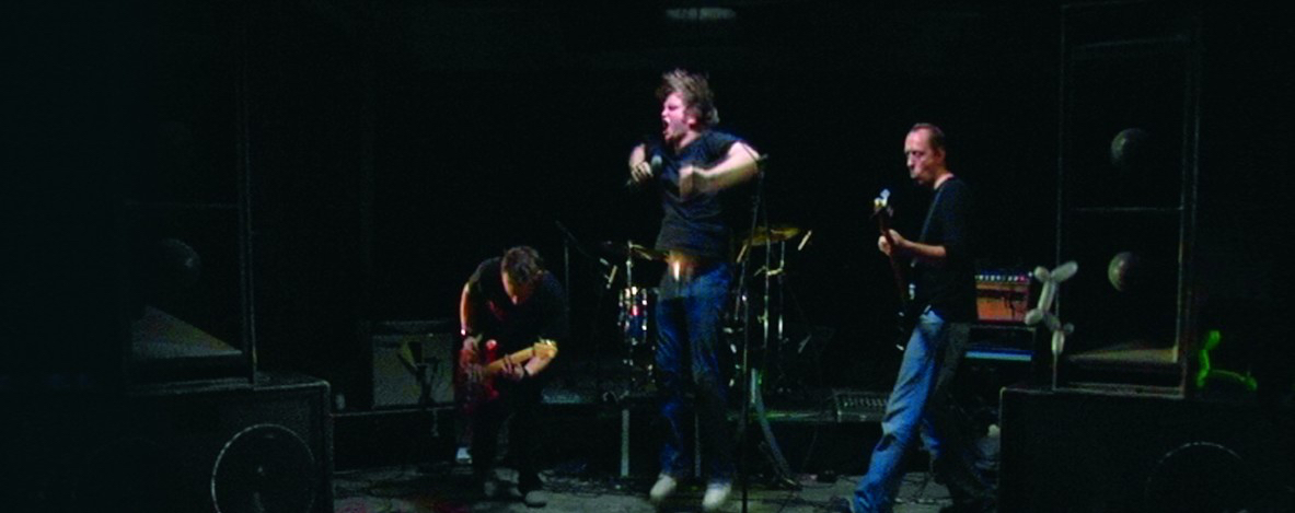 A still of 3 men performing in a band - two playing guitar and one person singing