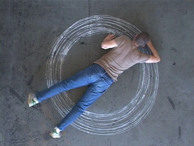 An aerial view of an artist stretching out on the floor with a piece of chalk. The artist draws concentric circles.
