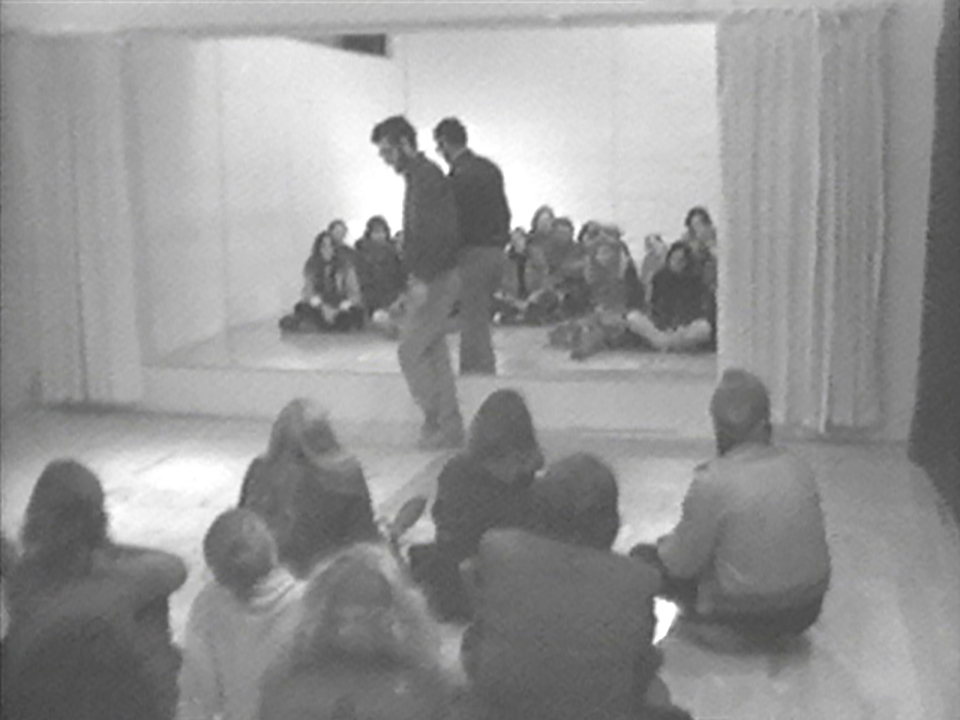 A small performance space with one man standing infront of a seated audience with a mirror in the back reflecting the stage.