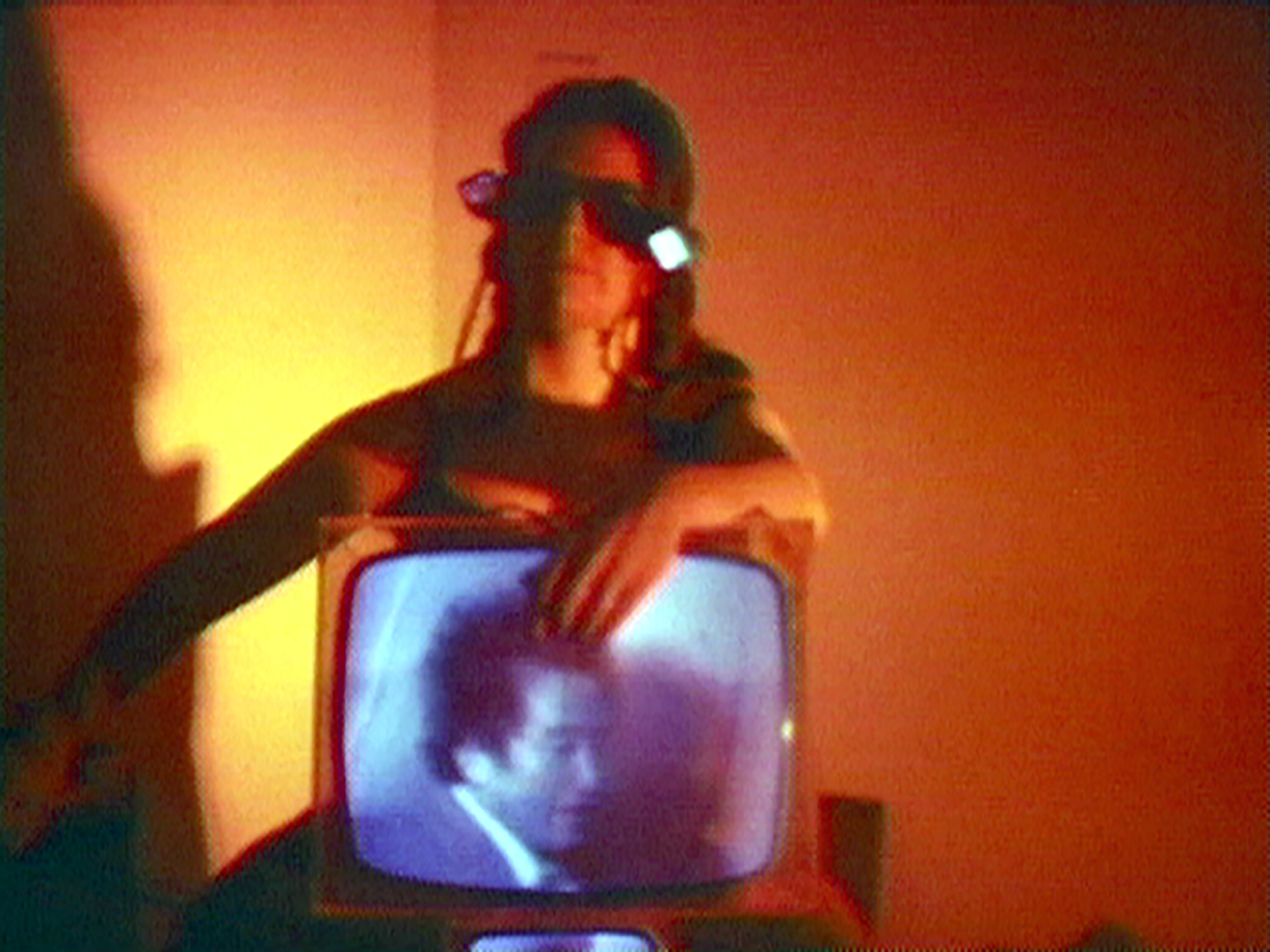Red hued image of a woman sitting with a retro television monitor in her lap. She is wearing glasses and the monitor depicts a man in a suit looking down.