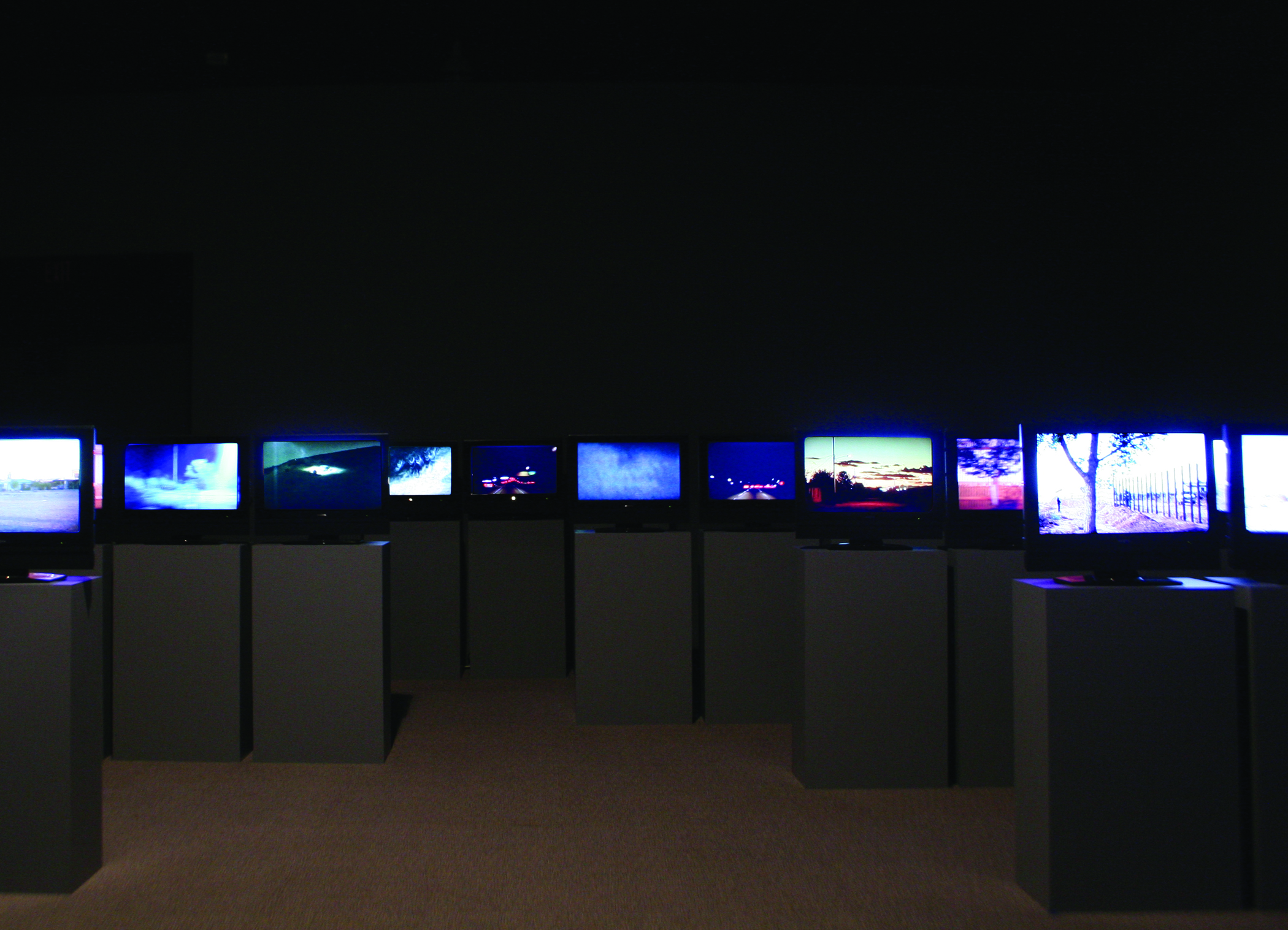 Many screens installed on podiums in a darkened gallery.