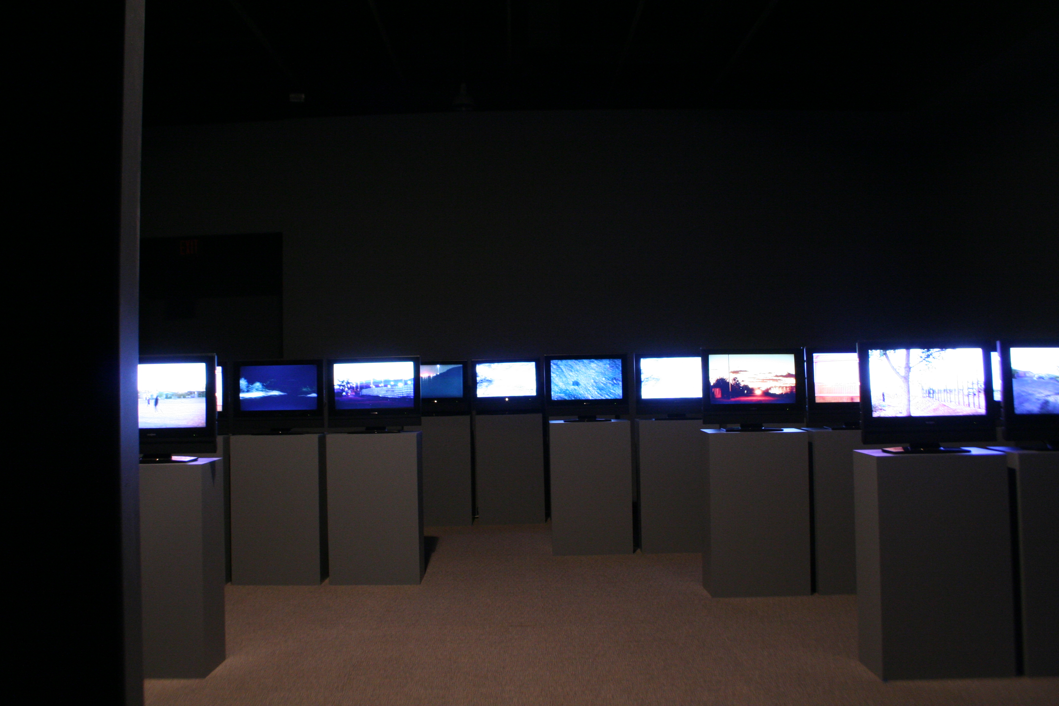 Many screens installed on podiums in a darkened gallery.