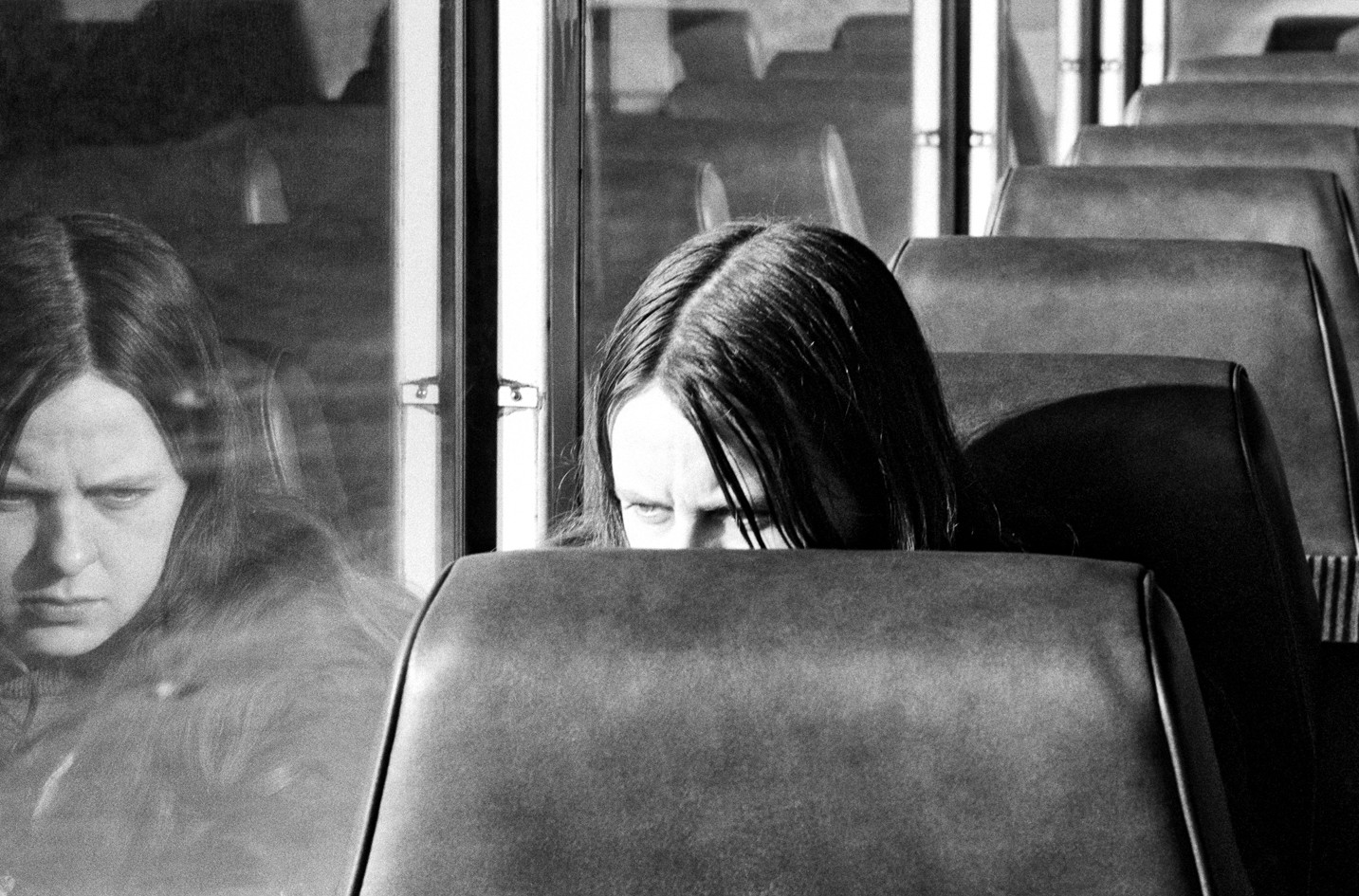 A girl on the bus looks at her own reflection in the window and is obscured by the seat in front of her.