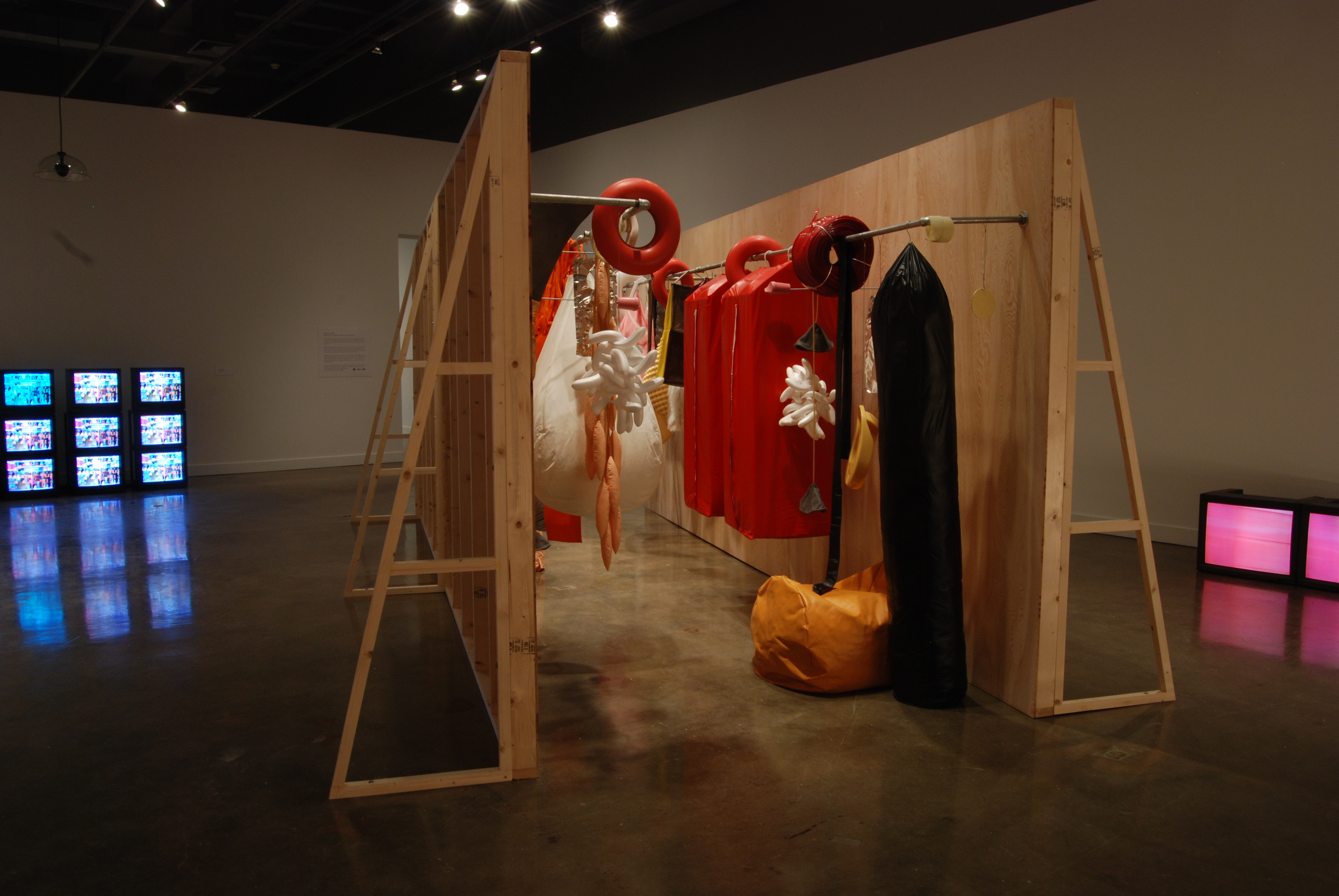 Two facing wood frame walls with orange, black, white objects hanging from racks. Stacked monitors in back and at right wall