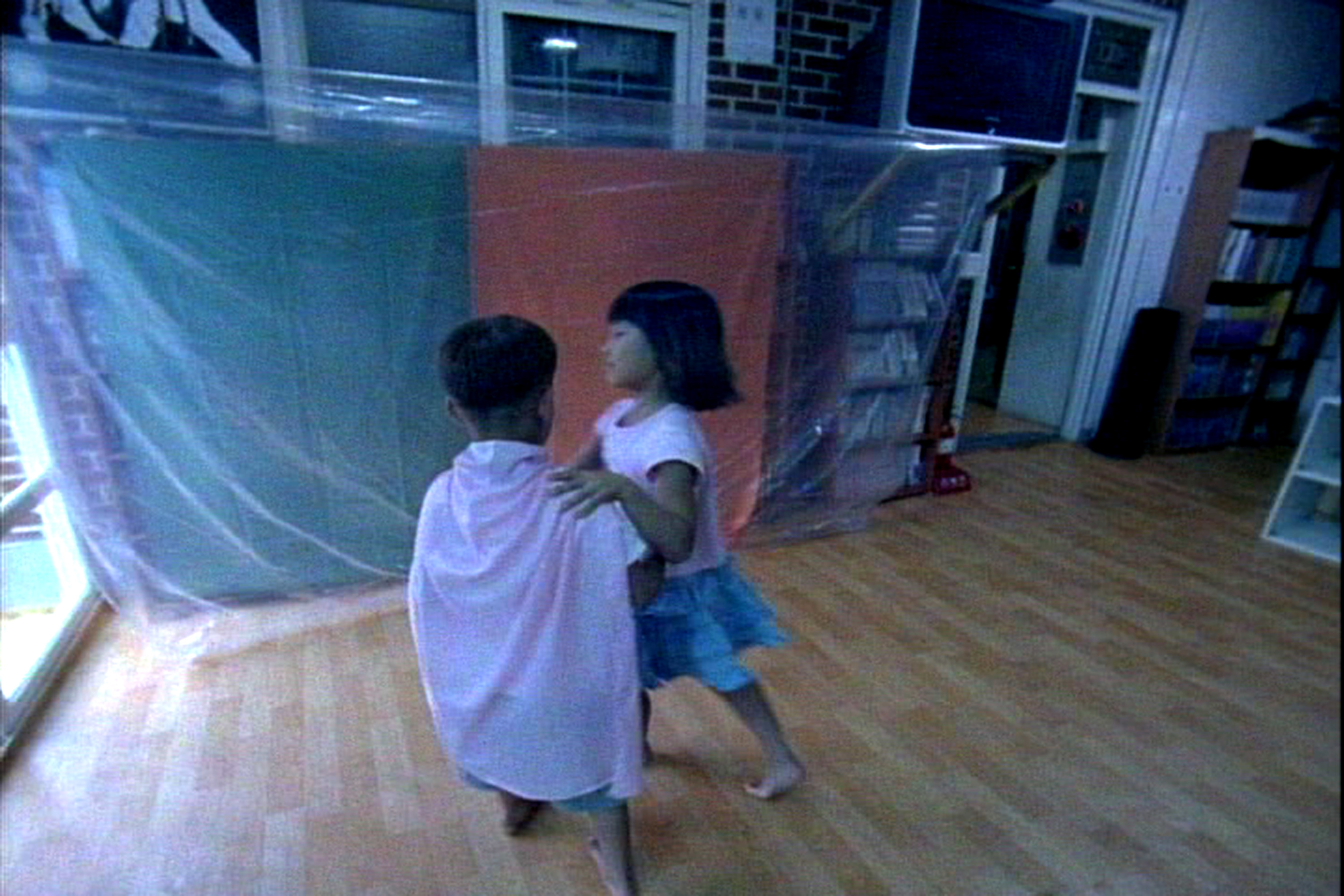 2 children dance bare-foot on a wooden floor in a room with bookshelves covered by green and orange cloth and clear plastic.
