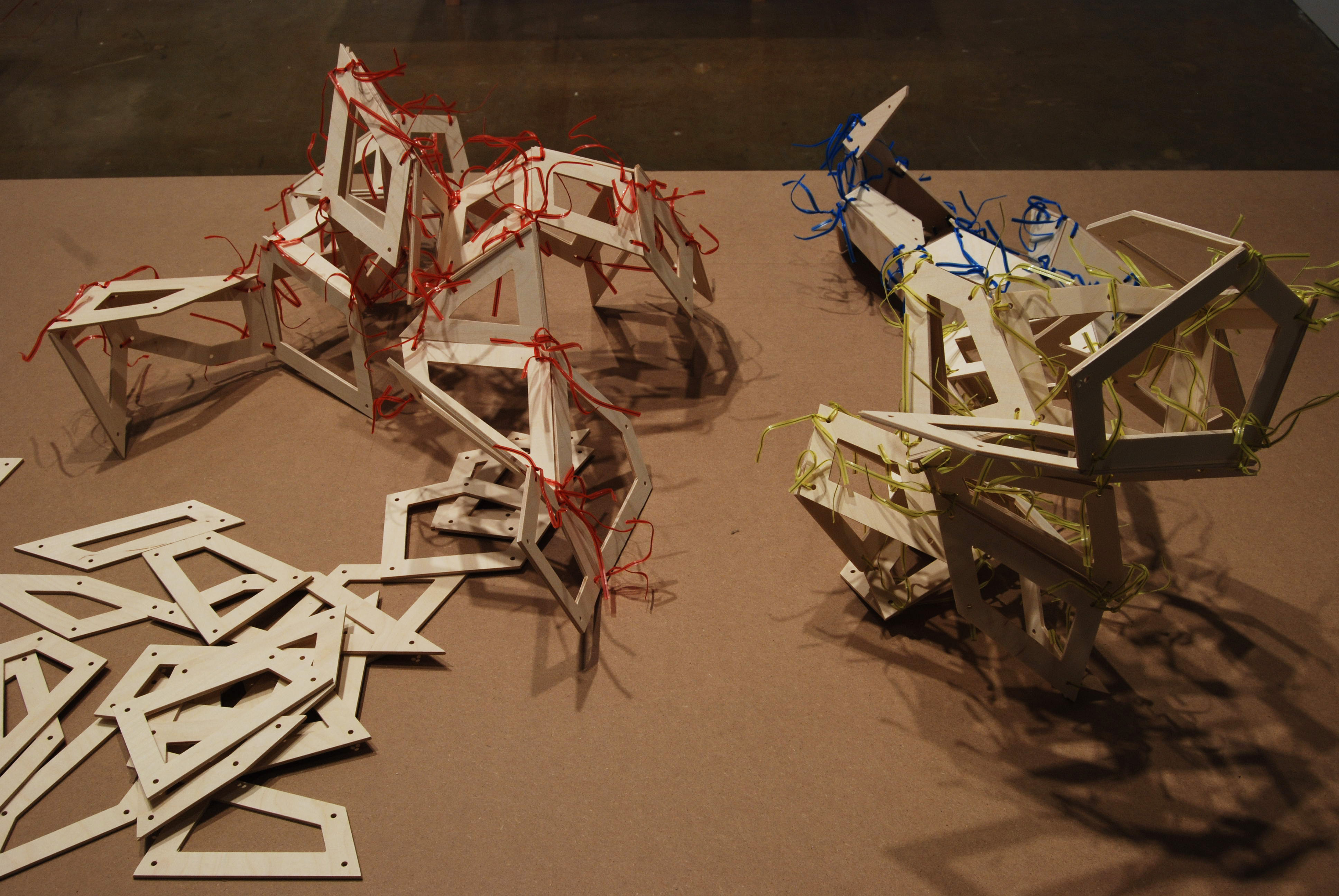 Small, perforated, flat geometric wood panels connected by red, yellow and blue ties form 3-dimensional tabletop sculptures.