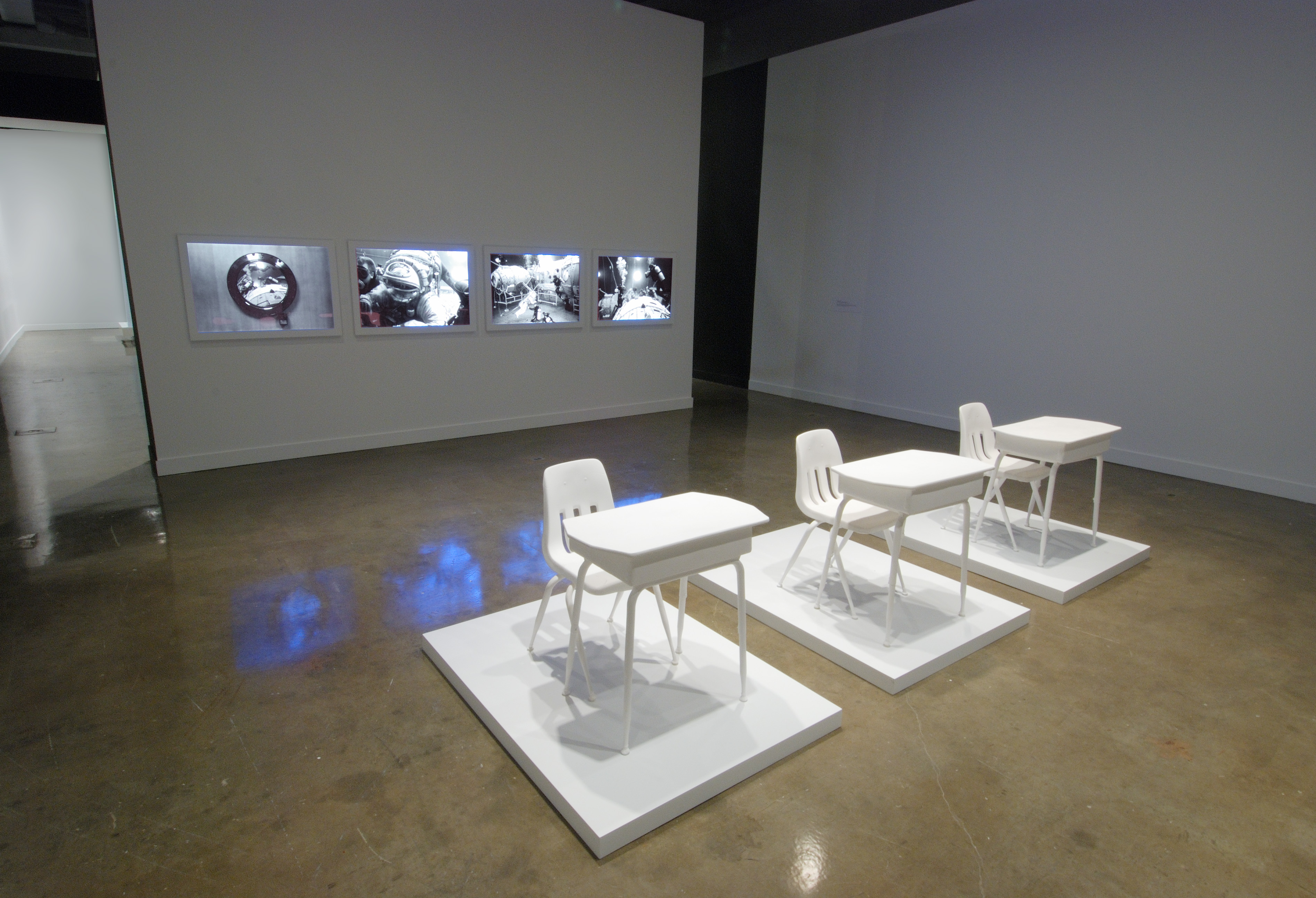 3 sets of white, school desks and chairs each on a white platform, before a gray wall mounted with 4 monitors