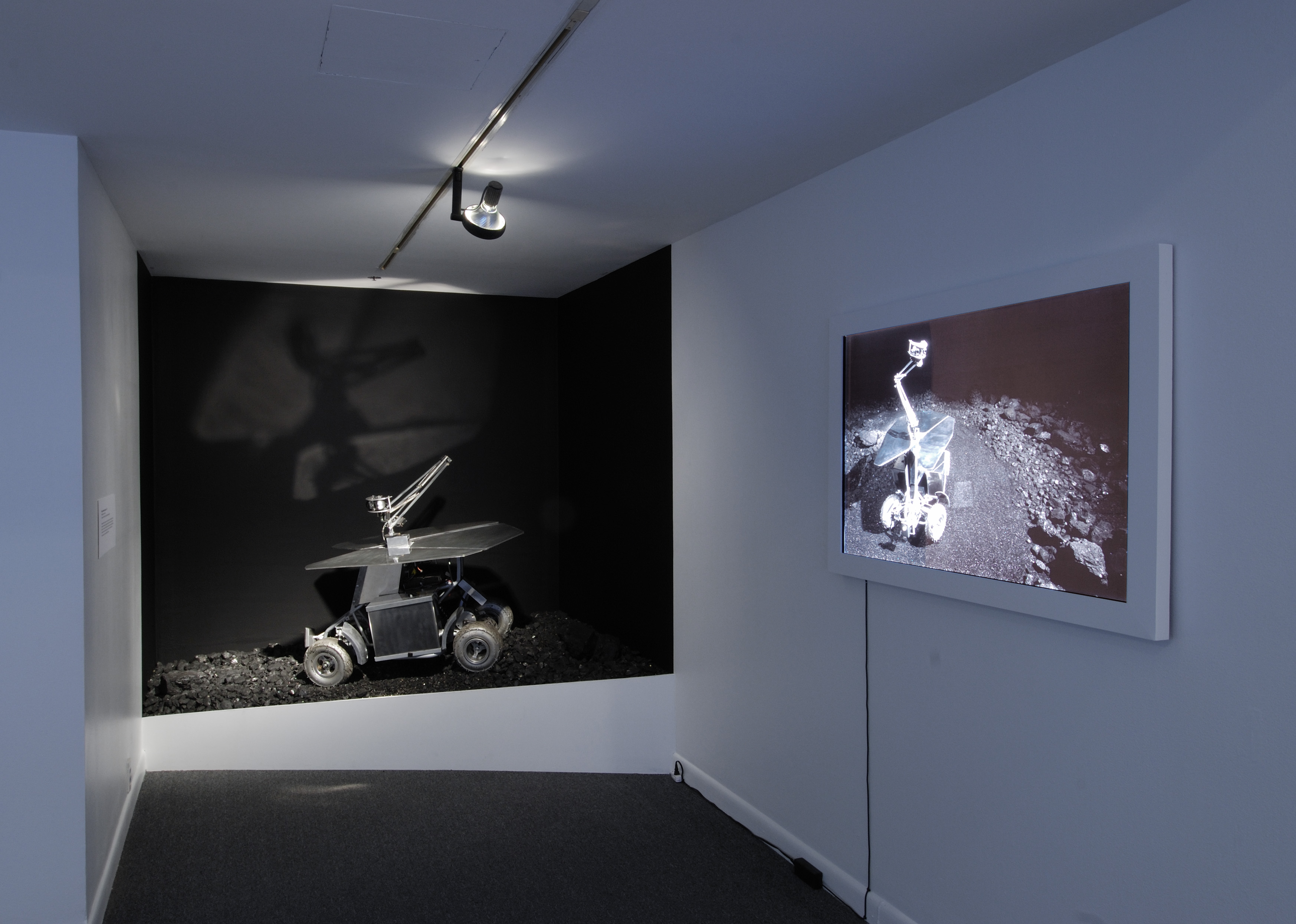Gray rover vehicle on slanted, coal-covered black platform next to wall-mounted monitor showing rover on rocky terrain