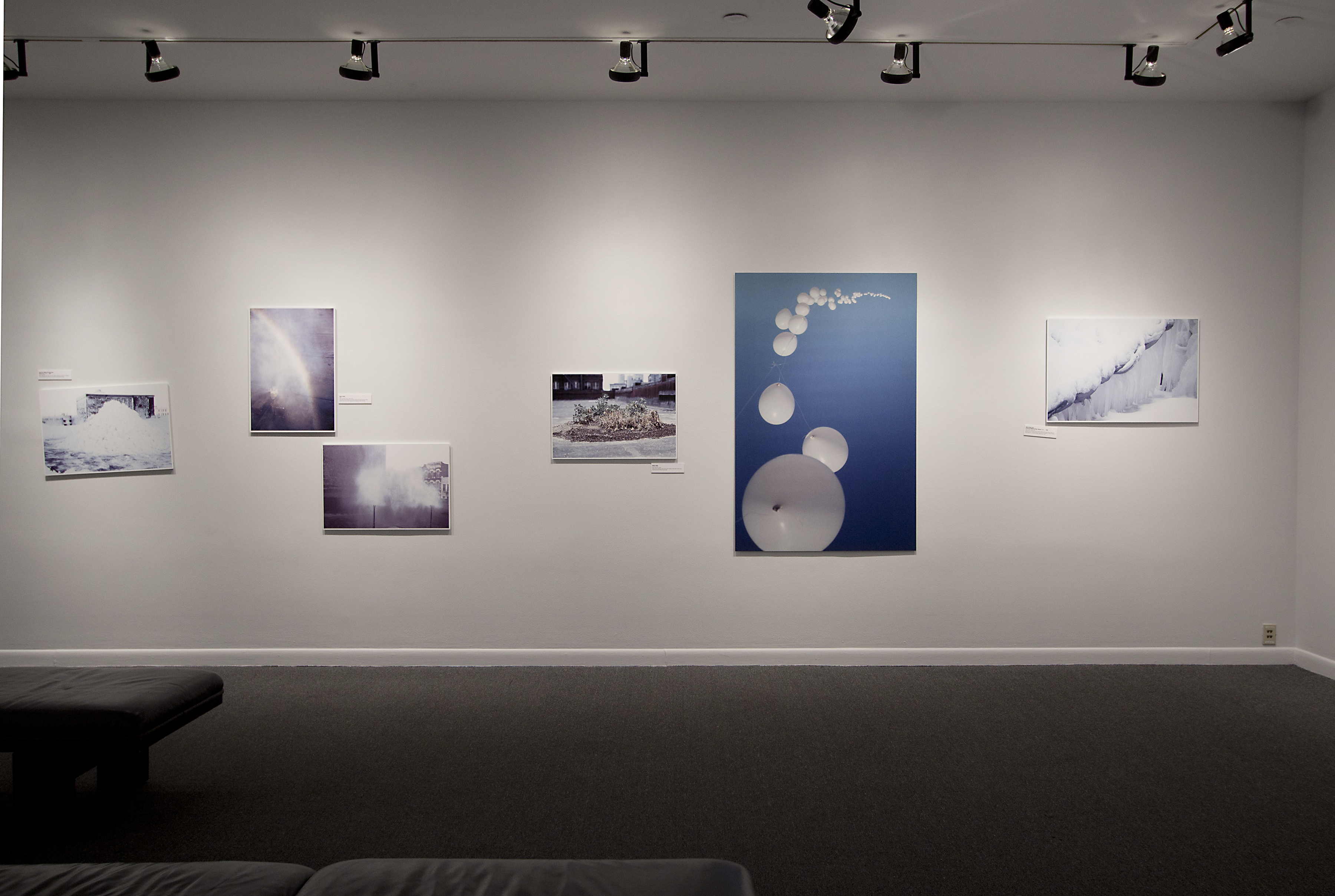 A gallery view of color photographs mounted at varying heights along a wall and lit from above with two benches in front.