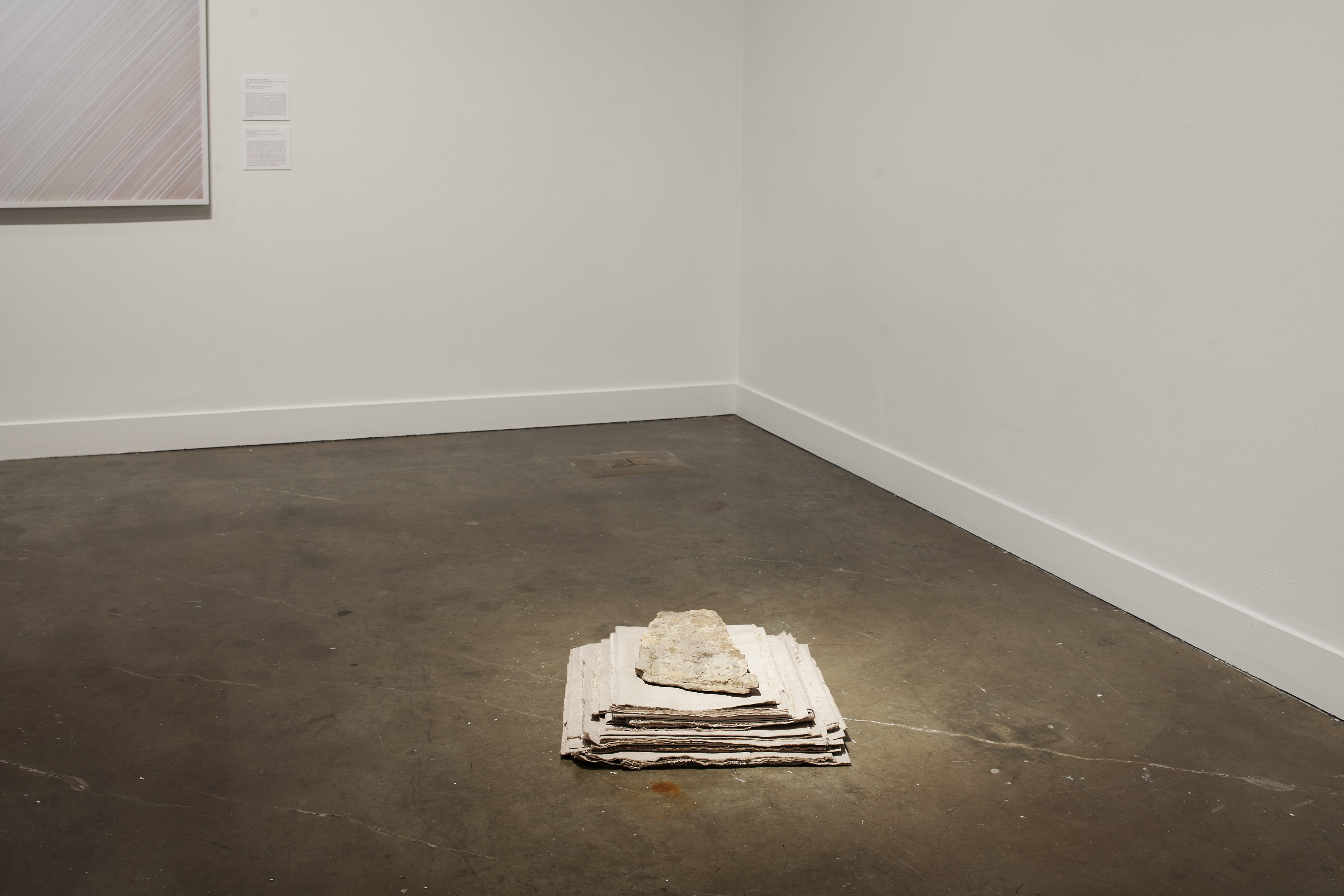A pile of folded fabric, largest at the bottom, with a flat rock on top and placed on the concrete floor near a corner